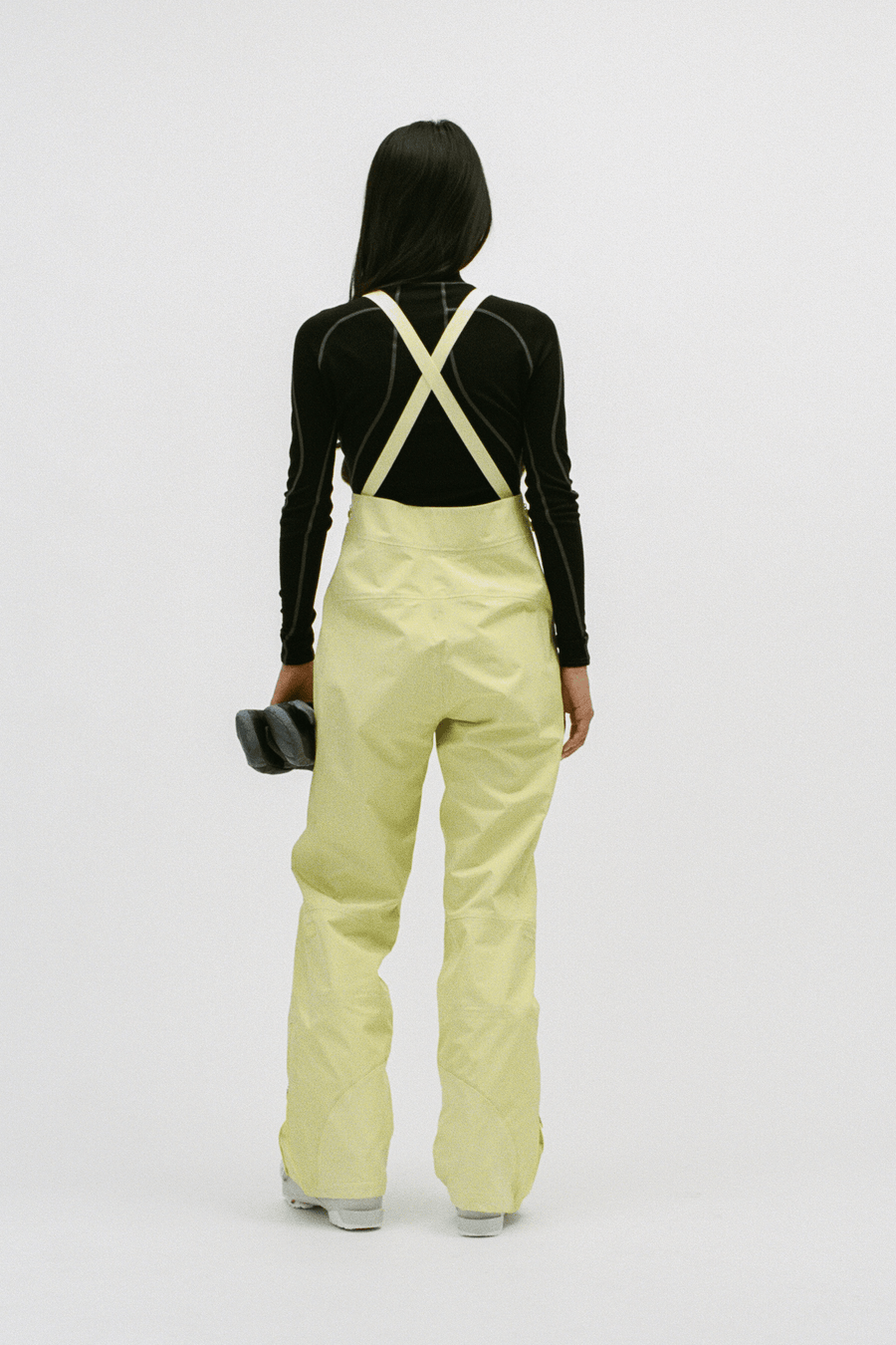 A model shown from the back wearing light yellow snow pants with cream-colored suspenders over a black long-sleeve top, holding a black ski boot against a plain white studio background.