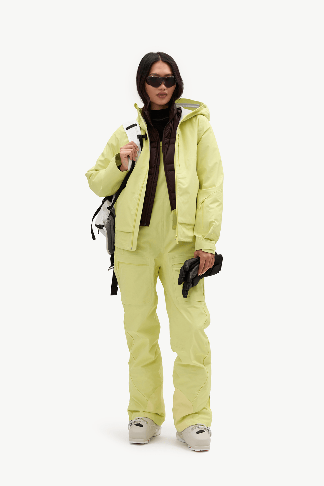 A model wearing a matching light yellow-green ski jacket and pants set, styled with a dark brown turtleneck, black sunglasses, black gloves, and beige ski boots against a plain white studio background.