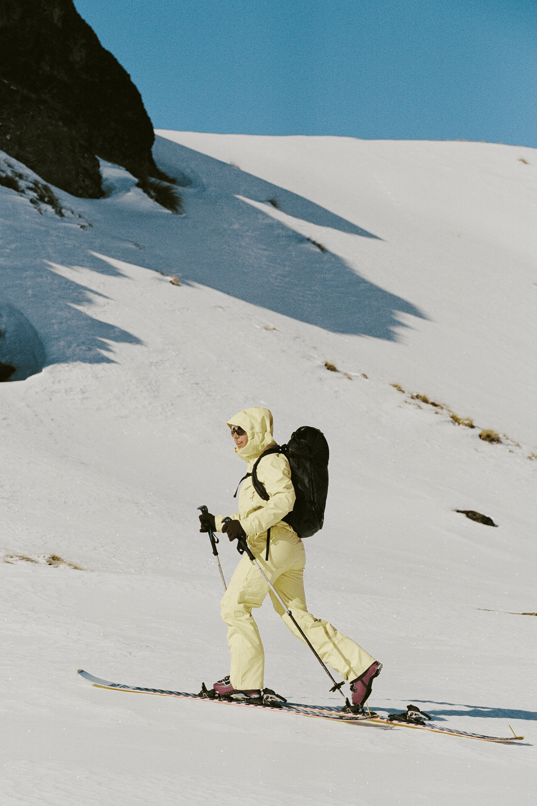 A skier in a pale yellow ski suit walks across a snowy mountain slope, carrying a black backpack and holding ski poles.