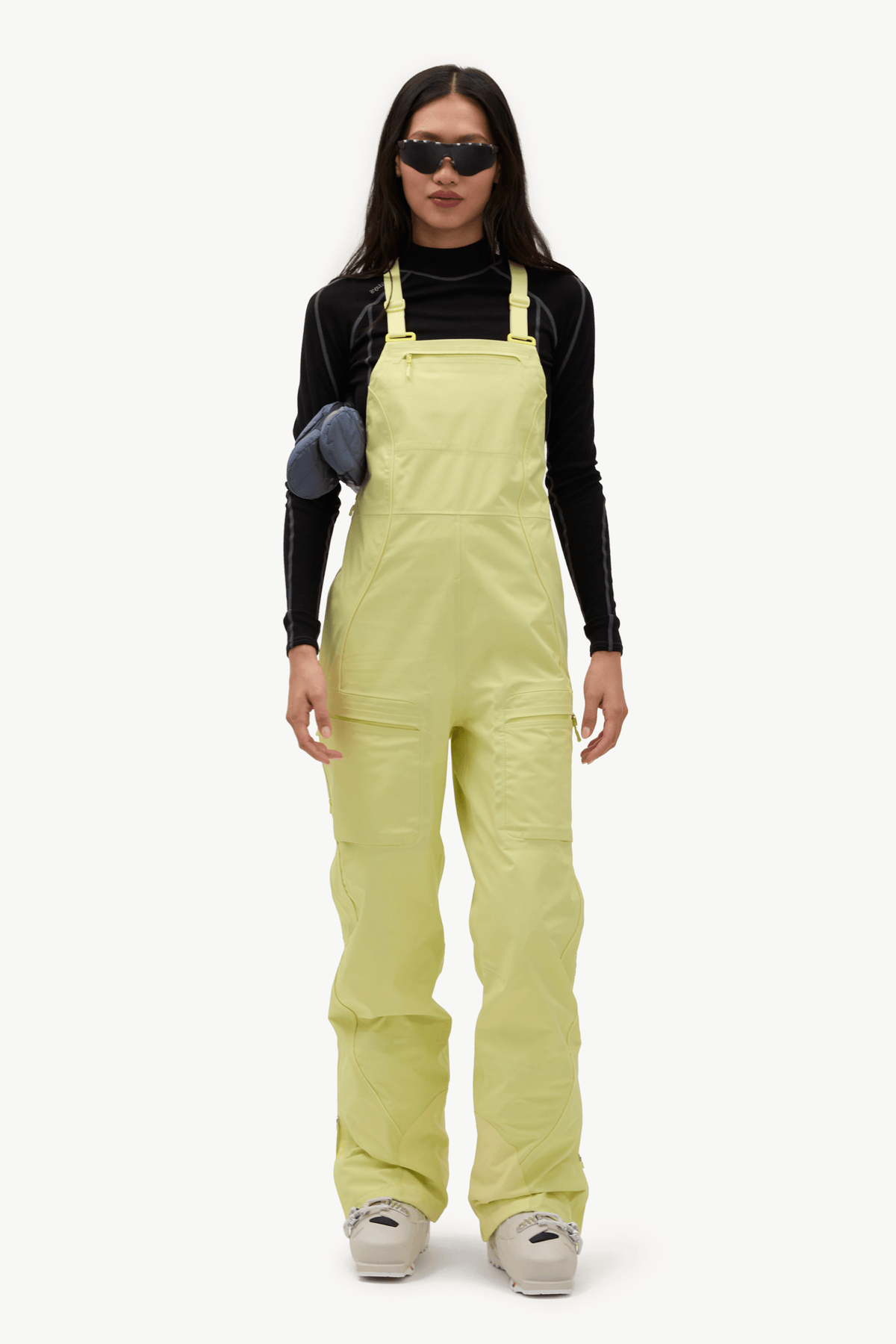 A model wearing light yellow-green snow bib overalls over a black long-sleeve top, styled with black sunglasses, a dark blue puffer jacket, and off-white sneakers, set against a plain white studio background.