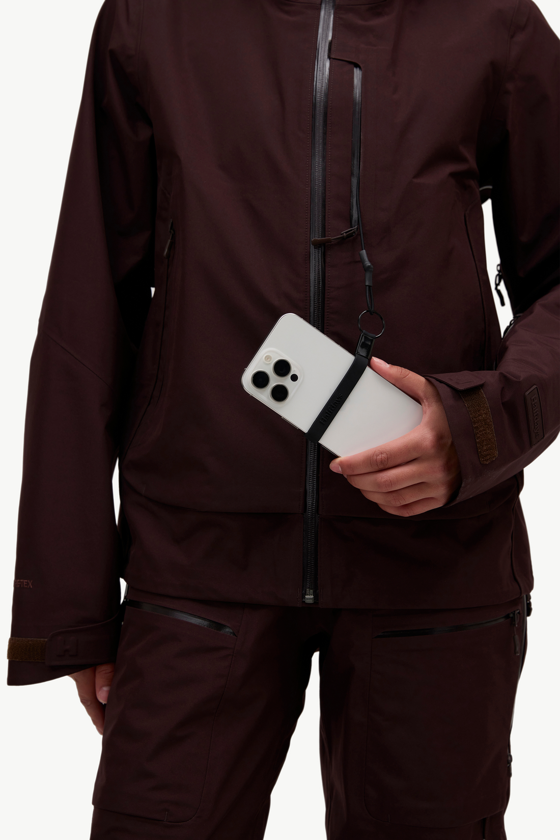 A close-up of a person wearing a dark brown technical outdoor jacket and matching pants, holding a white smartphone in a side pocket. The jacket has black zipper details and a small vertical brand patch on the sleeve.