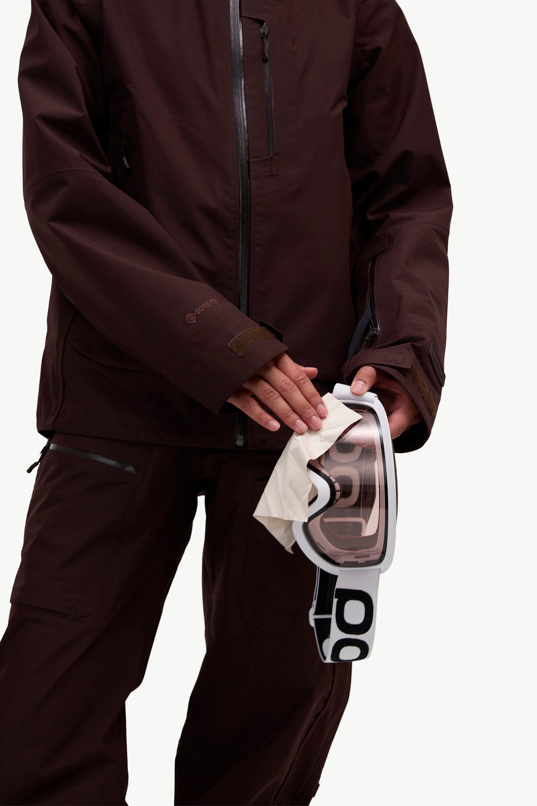 A person wearing a dark brown ski jacket and matching pants holds a pair of white and black goggles with a clear lens against a clean white background.