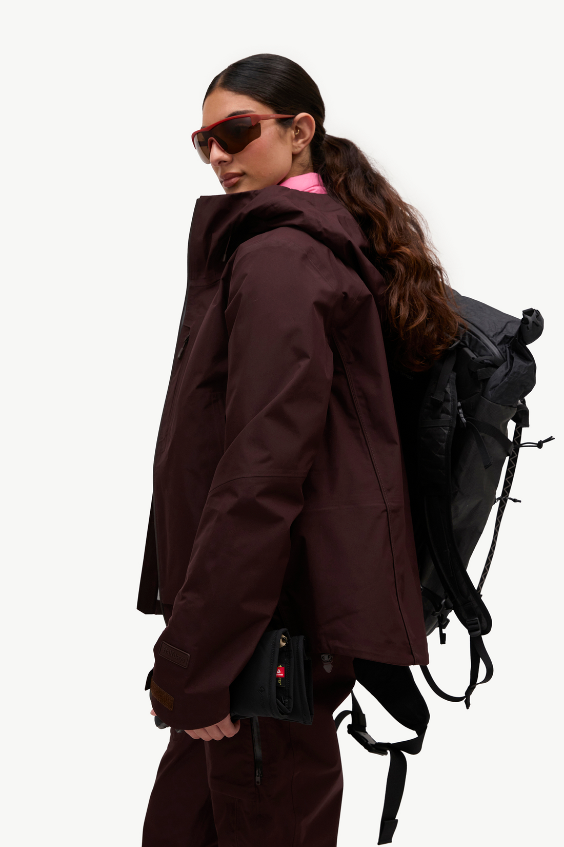 A woman in profile wearing a dark burgundy hooded jacket, matching pants, a black backpack, and red-framed sunglasses, photographed against a clean white studio background.