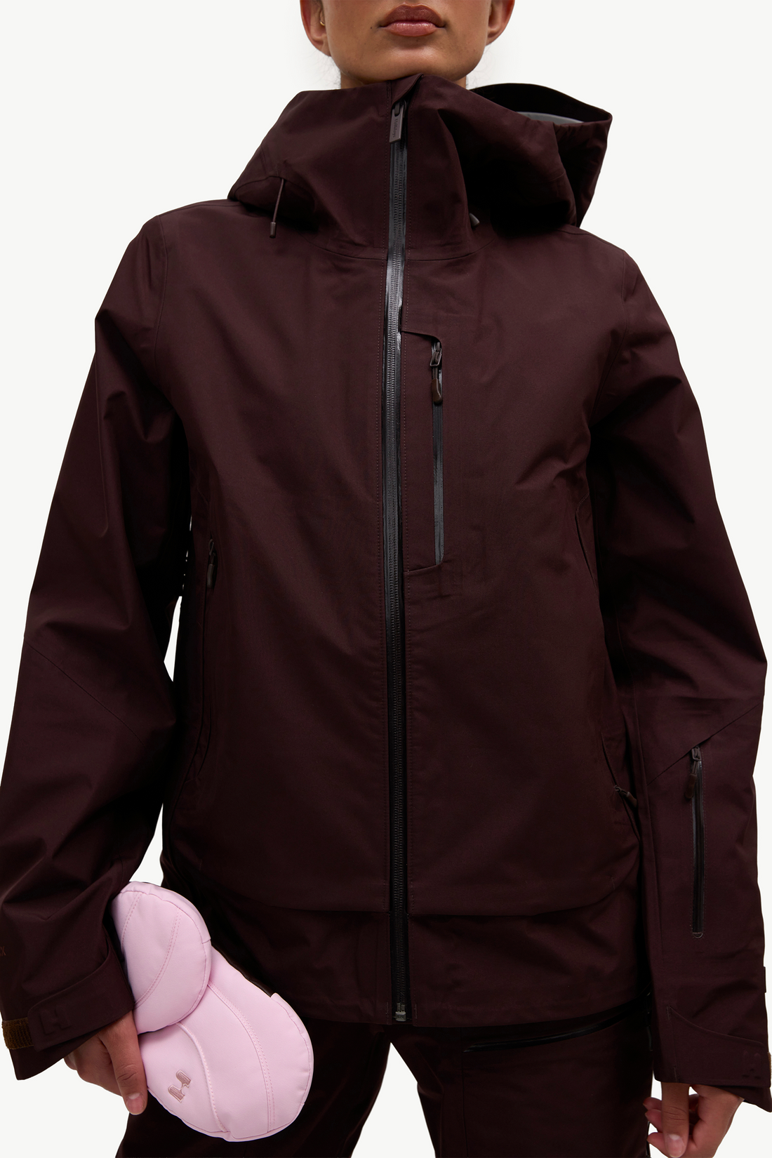 A person wearing a dark burgundy hooded ski jacket with black zippers, holding a light pink helmet against a plain white studio background.