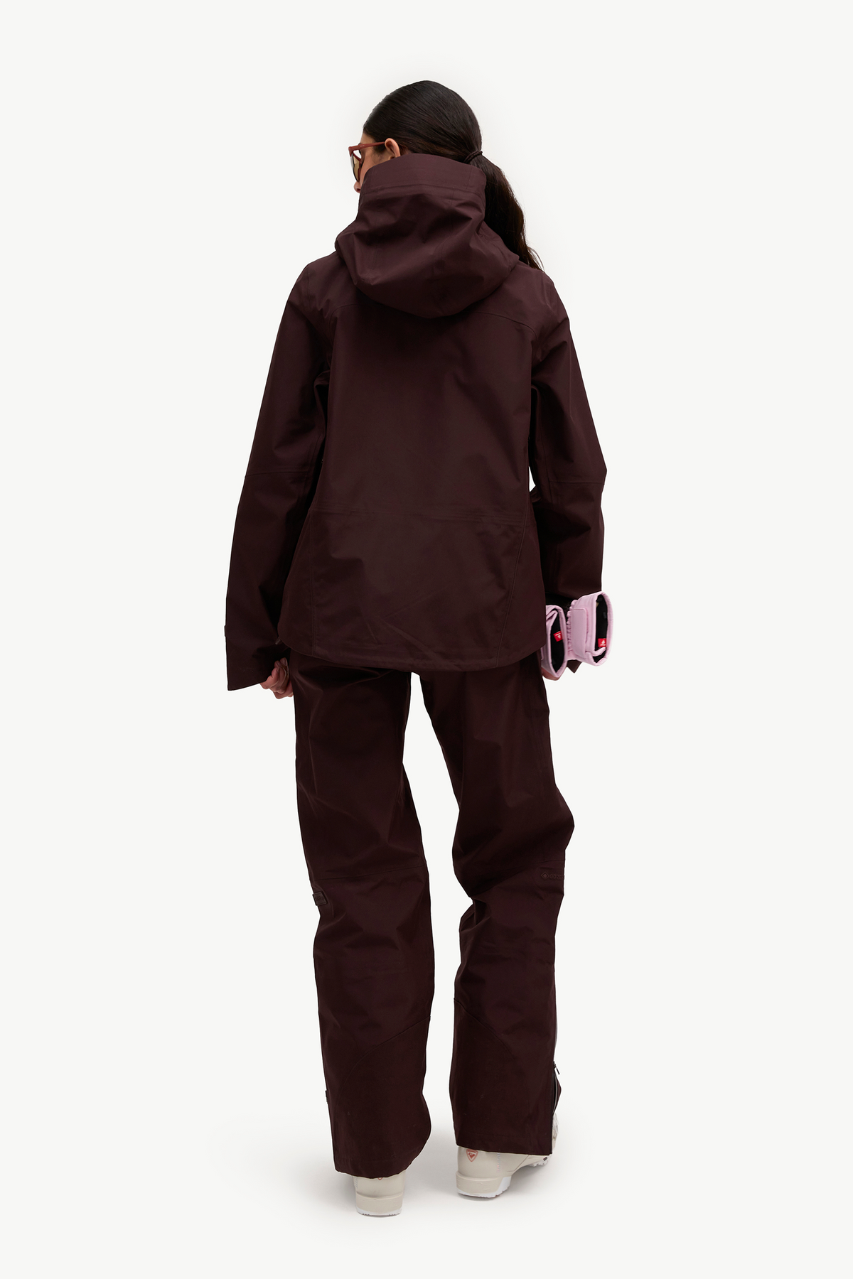 A model shown from the back wearing a matching dark plum-brown hooded jacket and pants set, holding a pink phone case against a clean white background.
