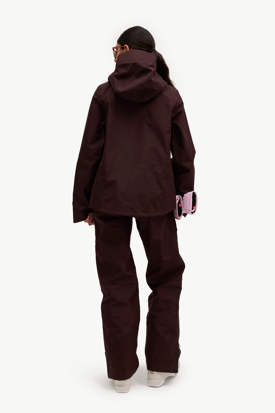 A model shown from the back wearing a matching dark plum-brown hooded jacket and pants set, holding a pink phone case against a clean white background.