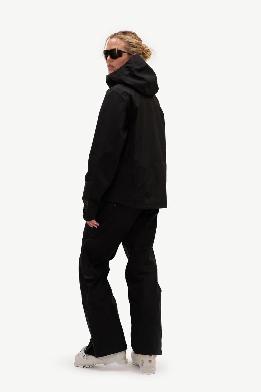A woman modeling a black hooded jacket and black snow pants, shown from the side against a plain white studio background.