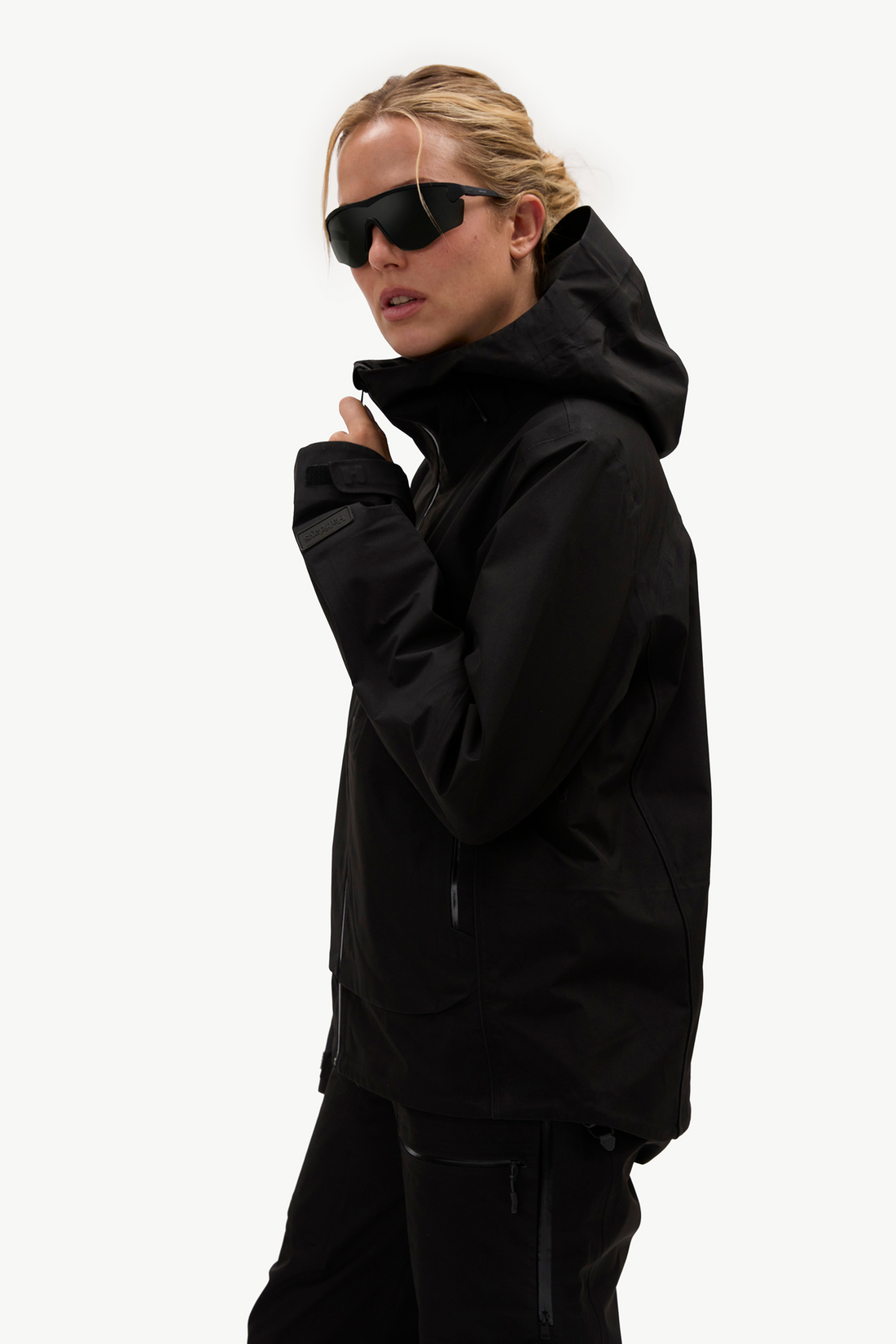 A woman in a black hooded ski jacket and black pants, wearing black sunglasses, shown in profile against a clean white studio background.