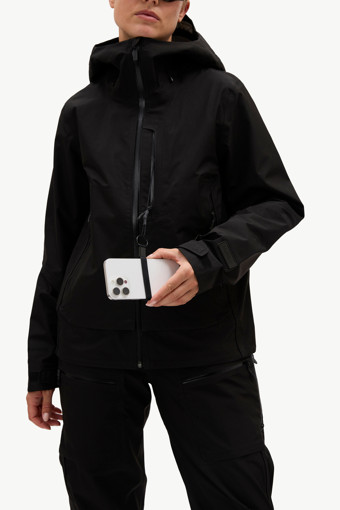 A model wearing a black hooded technical jacket and matching black pants, holding a white smartphone in front of the jacket pocket, against a clean white studio background.
