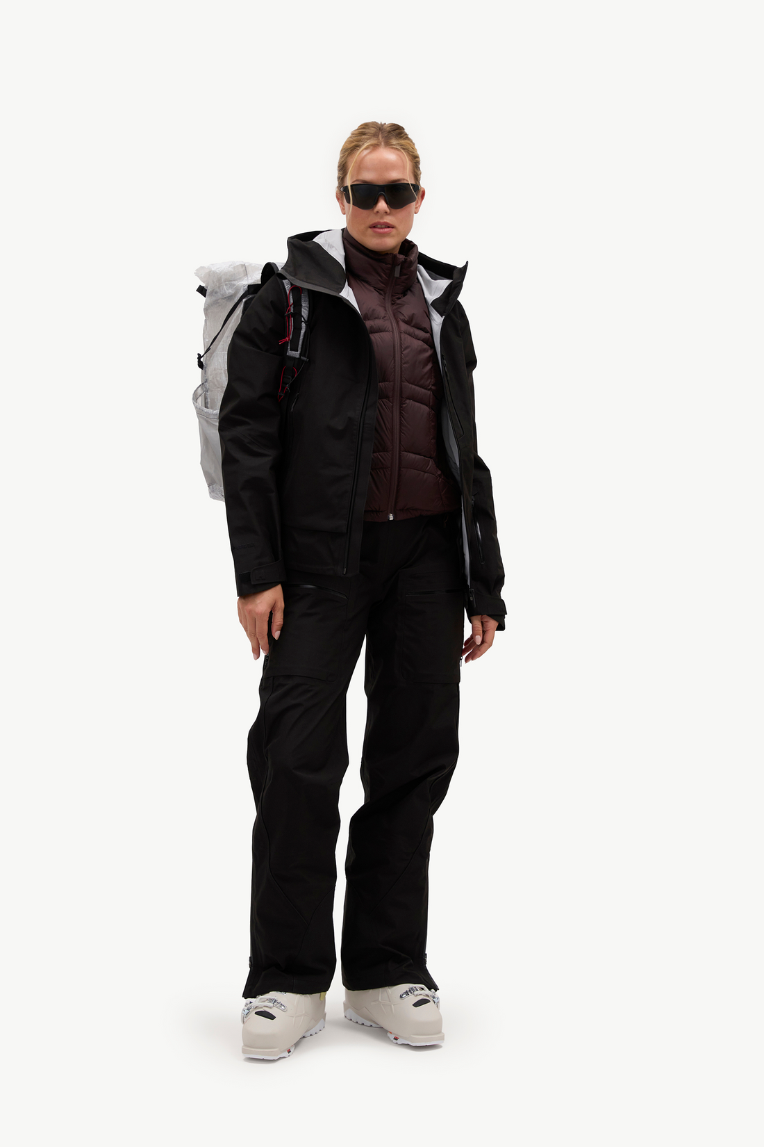 A model stands against a plain white studio background wearing a black ski jacket, black ski pants, white ski boots, black sunglasses, and a light gray backpack.