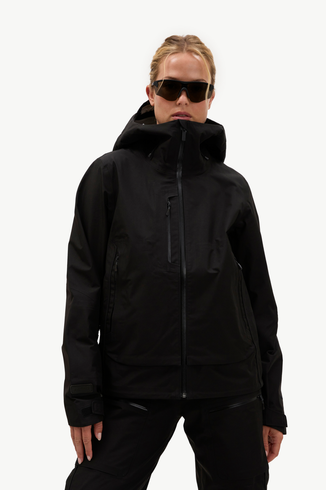 A model wearing a black hooded technical jacket and matching black pants, styled with black sunglasses, against a solid white studio background.