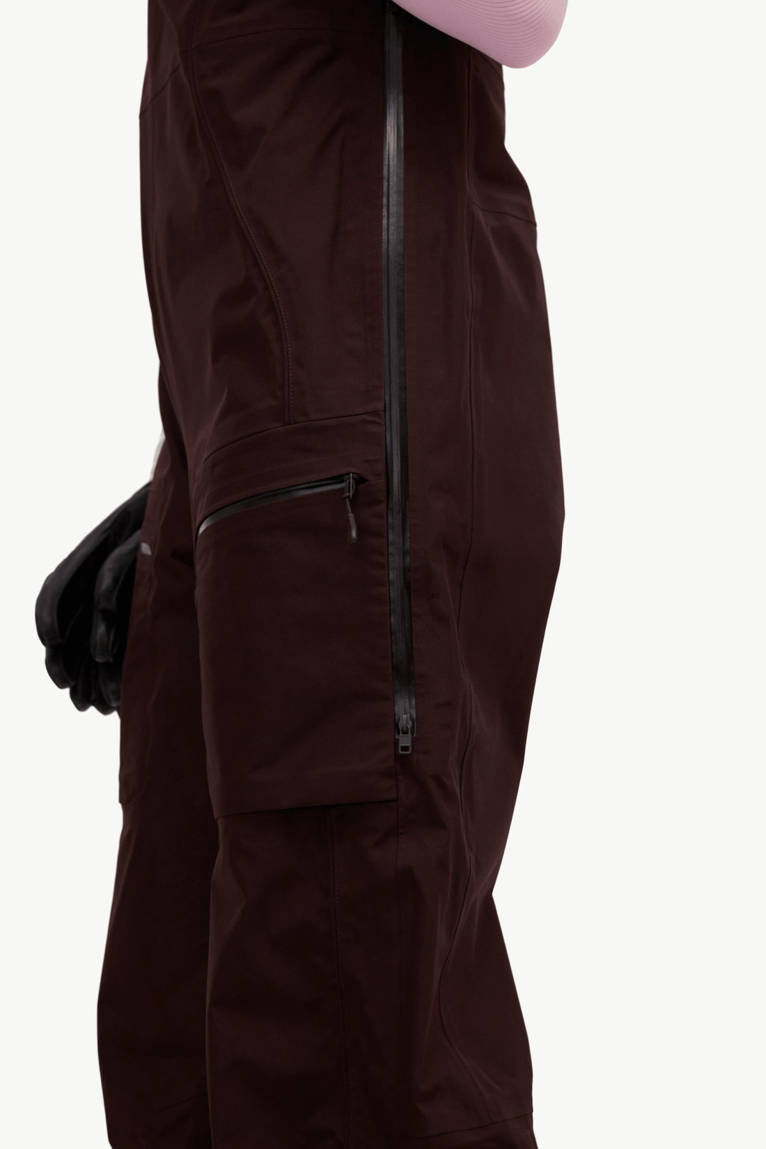 Close-up side view of dark brown snow pants with black zippers and a black glove, against a light gray background.