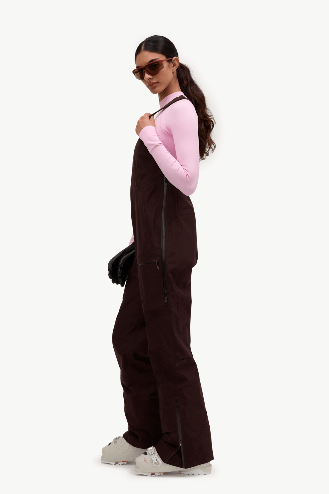 A model wearing dark brown corduroy snow bib overalls with black zippers, styled over a light pink long-sleeve top and white sneakers, set against a plain white studio background.