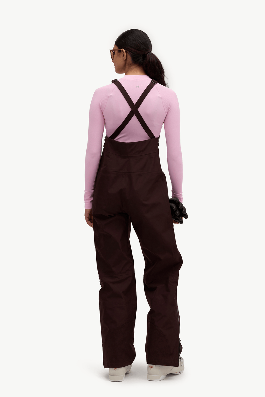 A model shown from the back wearing dark brown snow bib overalls over a light pink long-sleeve top, on a solid white background.