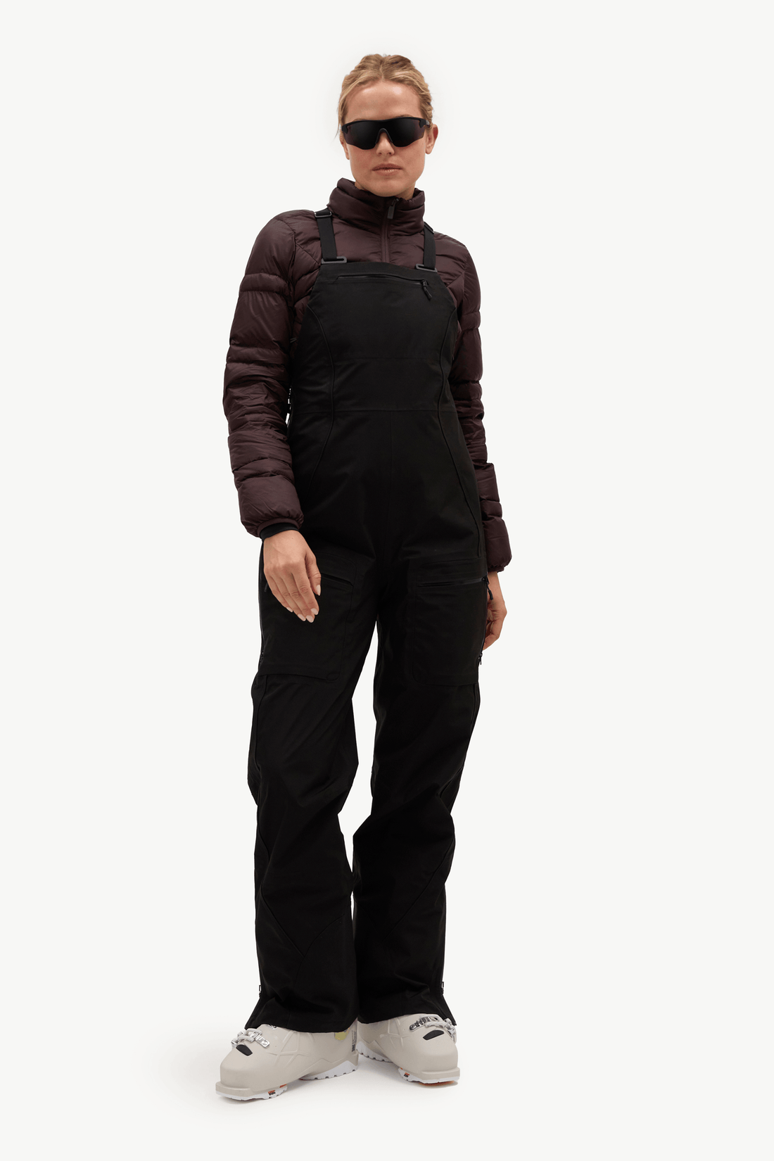 A model wearing black snow bib overalls, a dark brown puffer jacket, black sunglasses, and light gray ski boots, standing against a plain white studio background.