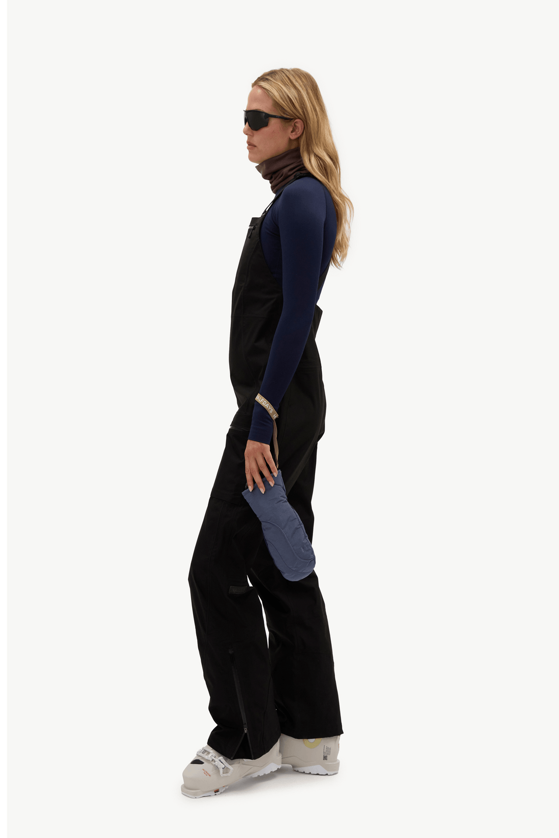A woman stands in profile wearing black snow bib overalls with a blue textured patch on the left knee, a navy long-sleeve top, a dark brown neck warmer, black sunglasses, and white ski boots against a plain white studio background.