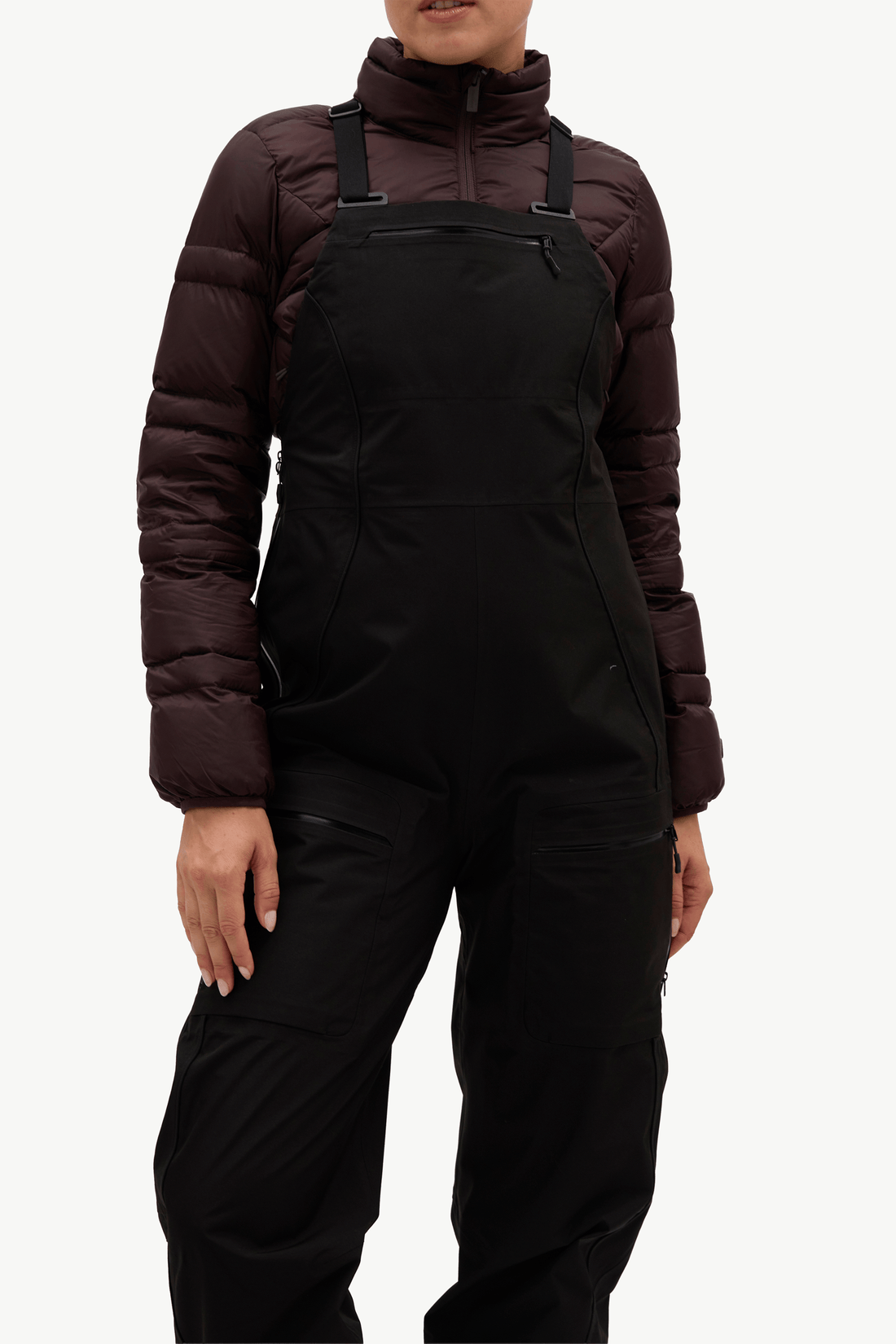 A model wearing black snow bib overalls with a dark brown puffer jacket, shown from the neck down against a plain white background.