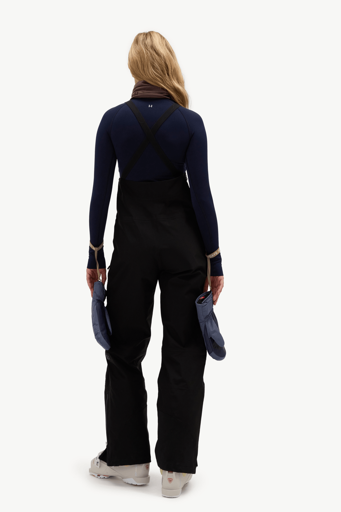 A woman viewed from the back wearing a dark navy long-sleeve top with black cross-back straps and black snow pants, holding blue gloves against a plain light background.