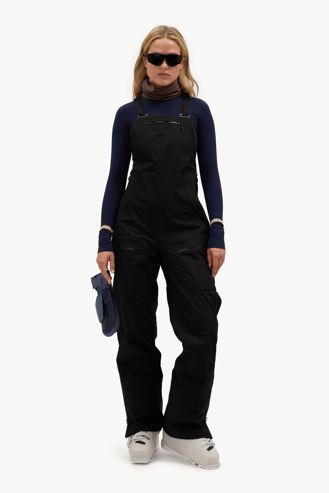 A model wearing black snow bib overalls over a navy blue long-sleeve top, brown neck gaiter, black sunglasses, and white ski boots, holding a pair of navy blue gloves against a plain white studio background.