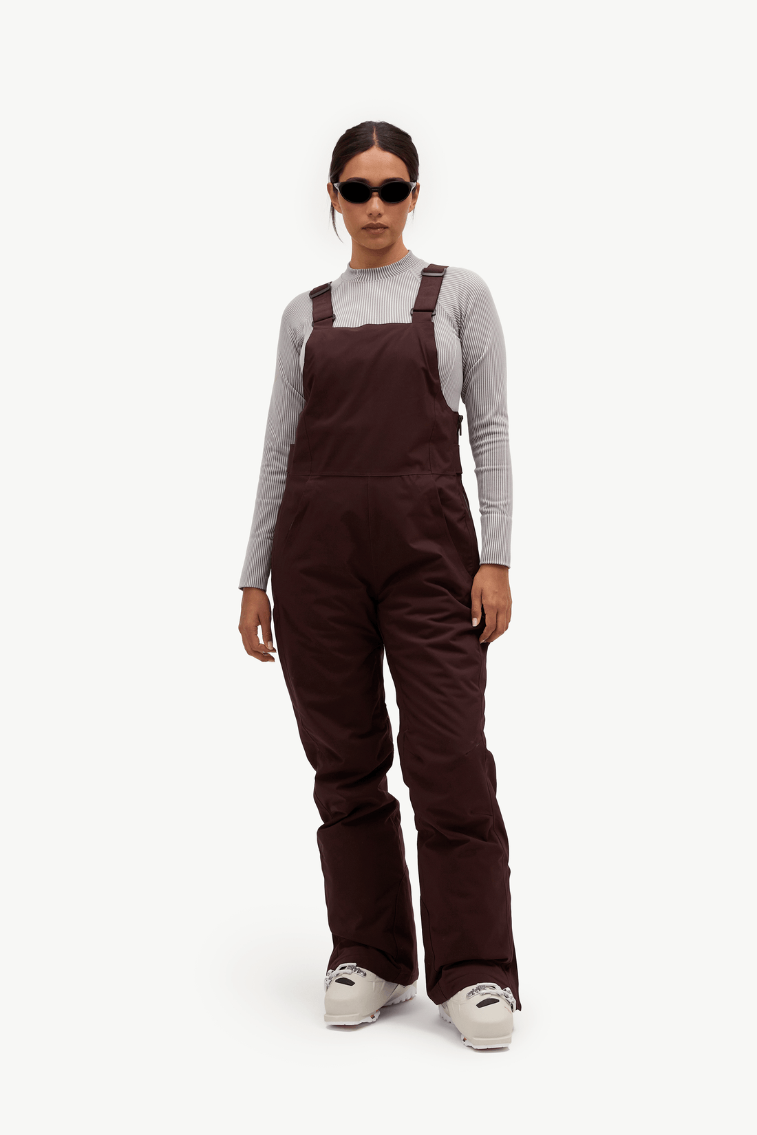A model wearing dark brown insulated bib overalls over a light gray long-sleeve shirt, styled with white sneakers and sunglasses, against a plain white studio background.