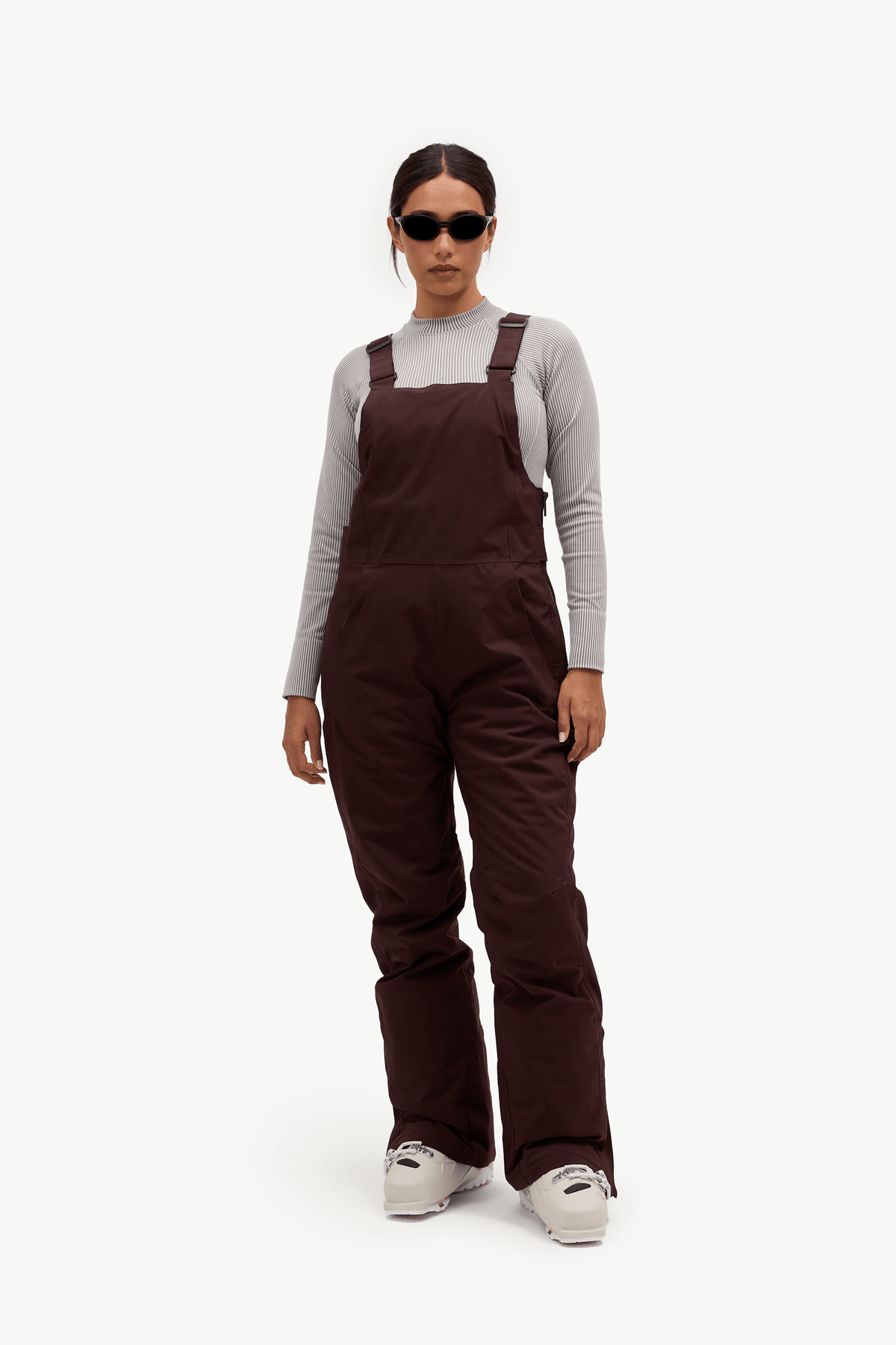 A model wearing dark brown insulated bib overalls over a light gray long-sleeve shirt, styled with white sneakers and sunglasses, against a plain white studio background.