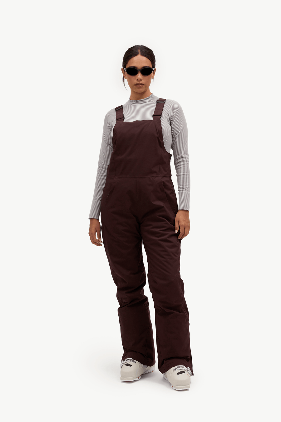 A model wearing dark brown insulated bib overalls over a light gray long-sleeve shirt, styled with white sneakers and sunglasses, against a plain white studio background.