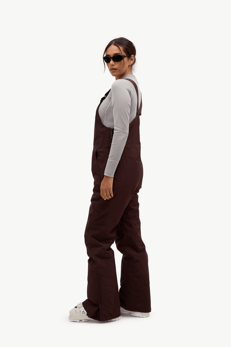 A model wearing dark brown wide-leg bib overalls over a light gray long-sleeve shirt, styled with white sneakers and sunglasses, photographed against a solid white background.