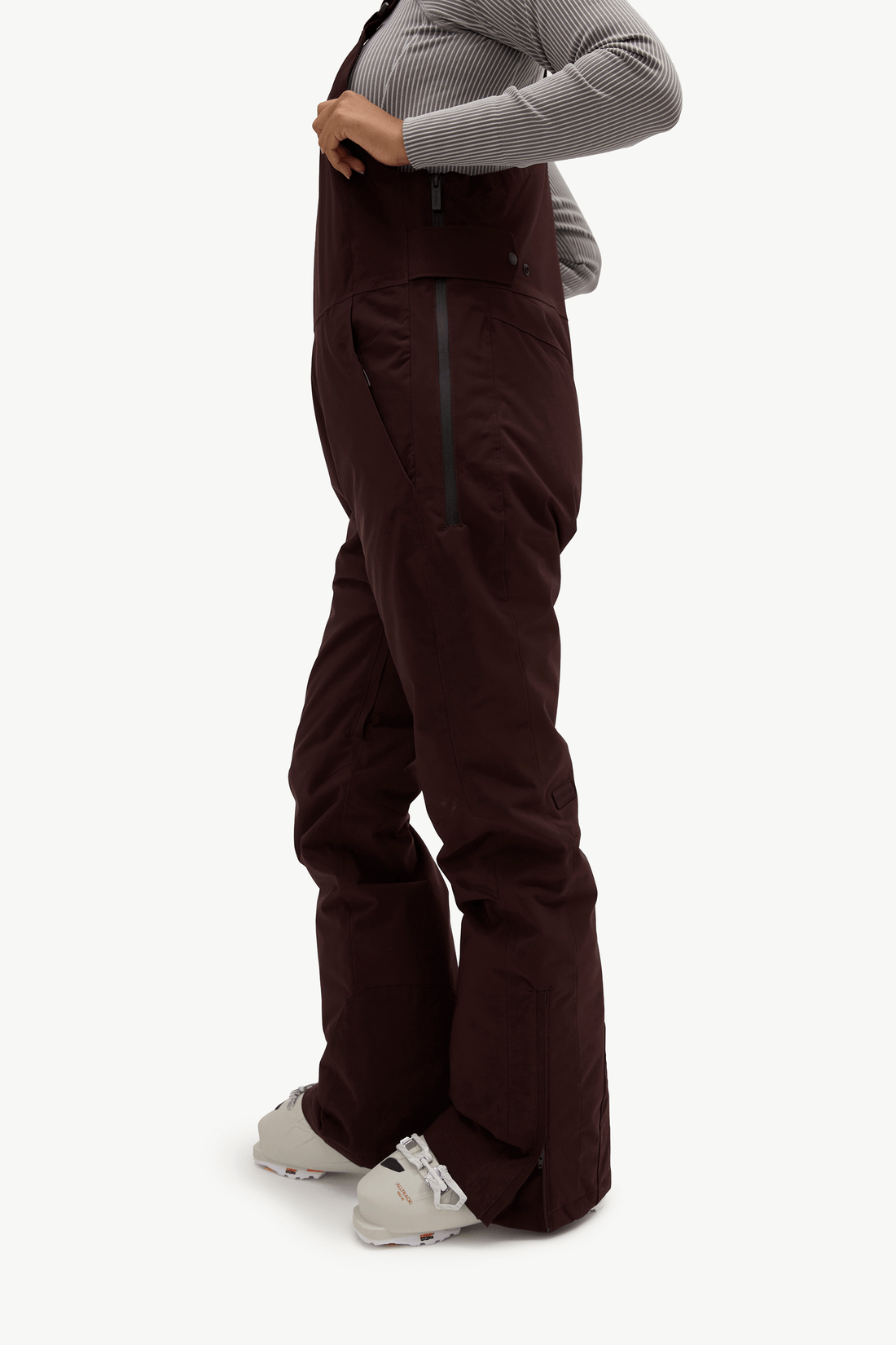A person wearing dark brown snow bib pants with a gray ribbed long-sleeve top and white sneakers, shown from the waist down against a plain light background.