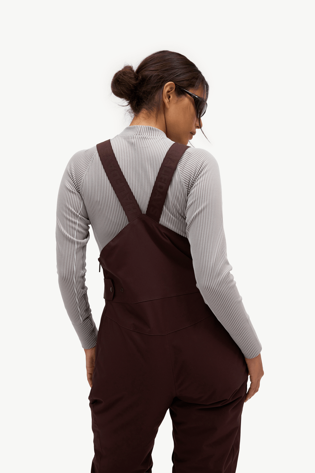 A woman shown from the back wearing dark brown overall-style bib overalls over a light gray ribbed long-sleeve top, photographed against a clean white studio background.