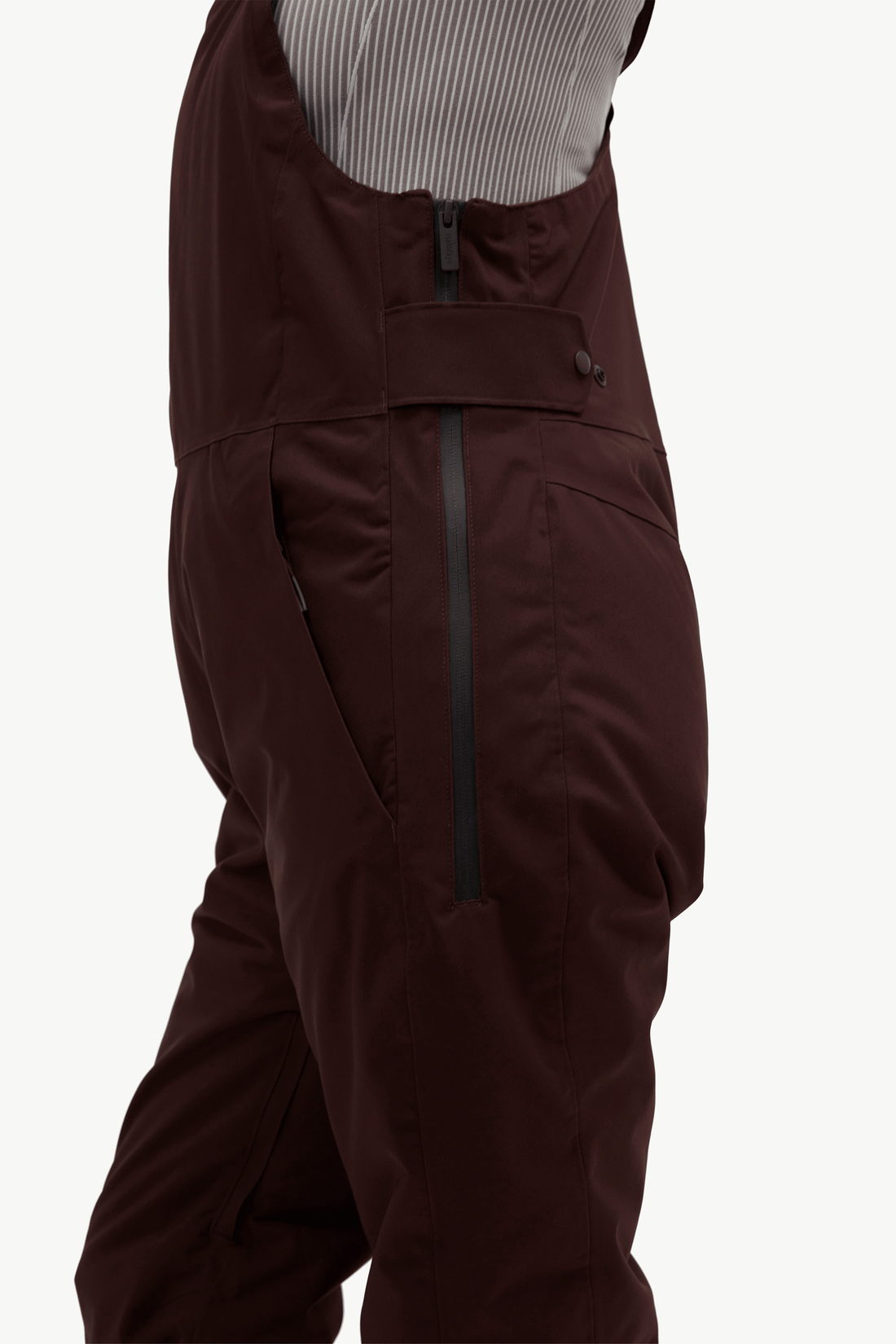 Close-up of dark burgundy insulated bib overalls with a vertical zipper and side pocket, worn over a striped shirt against a light gray background.