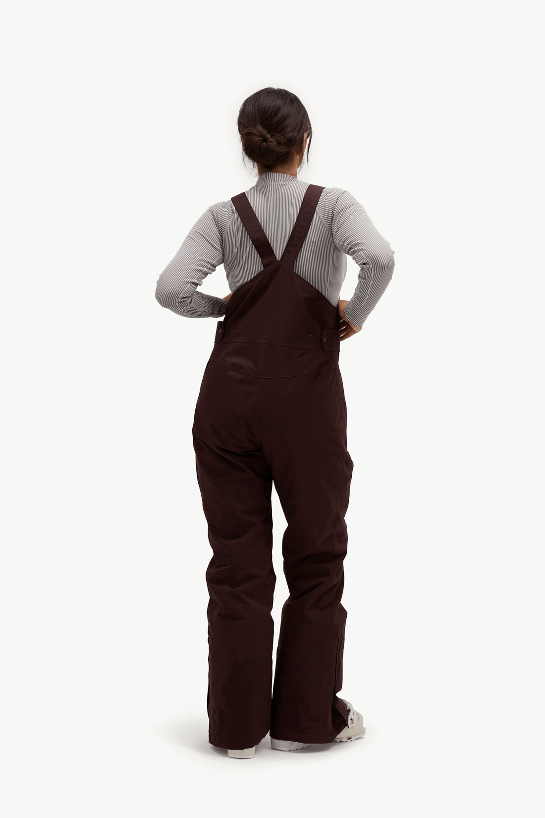 A person shown from the back wearing dark brown bib overalls over a light gray long-sleeve top, standing against a plain light gray studio background.
