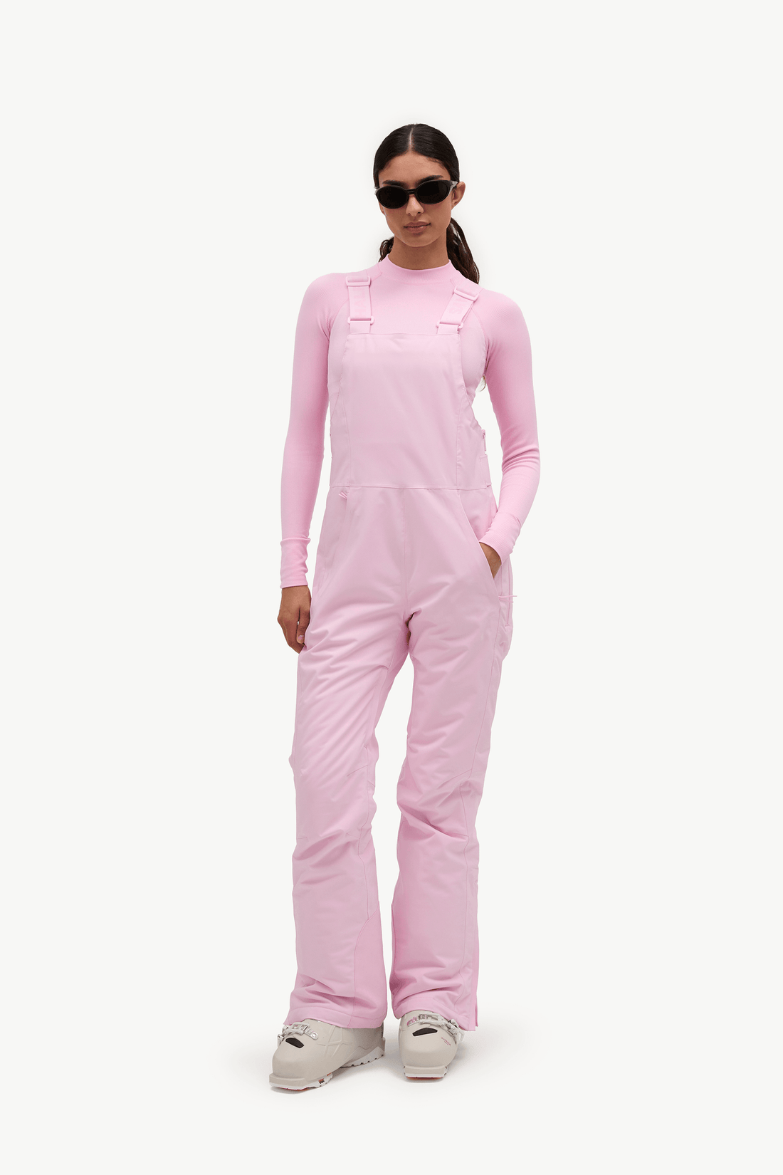 A model wearing a light pink long-sleeve snow bib jumpsuit with adjustable shoulder straps, styled with black sunglasses and light gray snowboard boots, set against a plain white studio background.