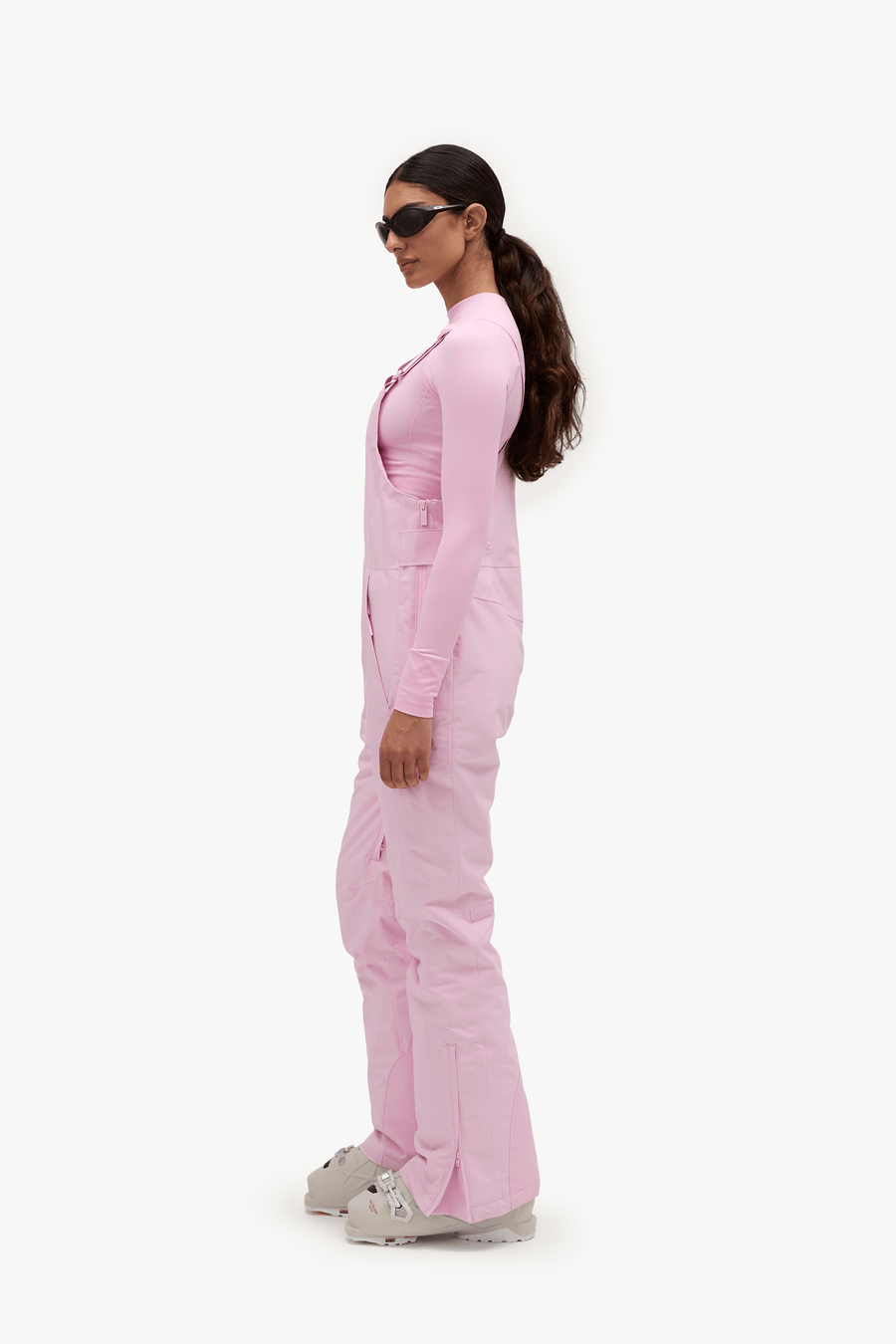 A woman in a side profile wearing a light pink long-sleeve scrub top and matching scrub pants, styled with white Croc sneakers and black sunglasses, against a plain white background.