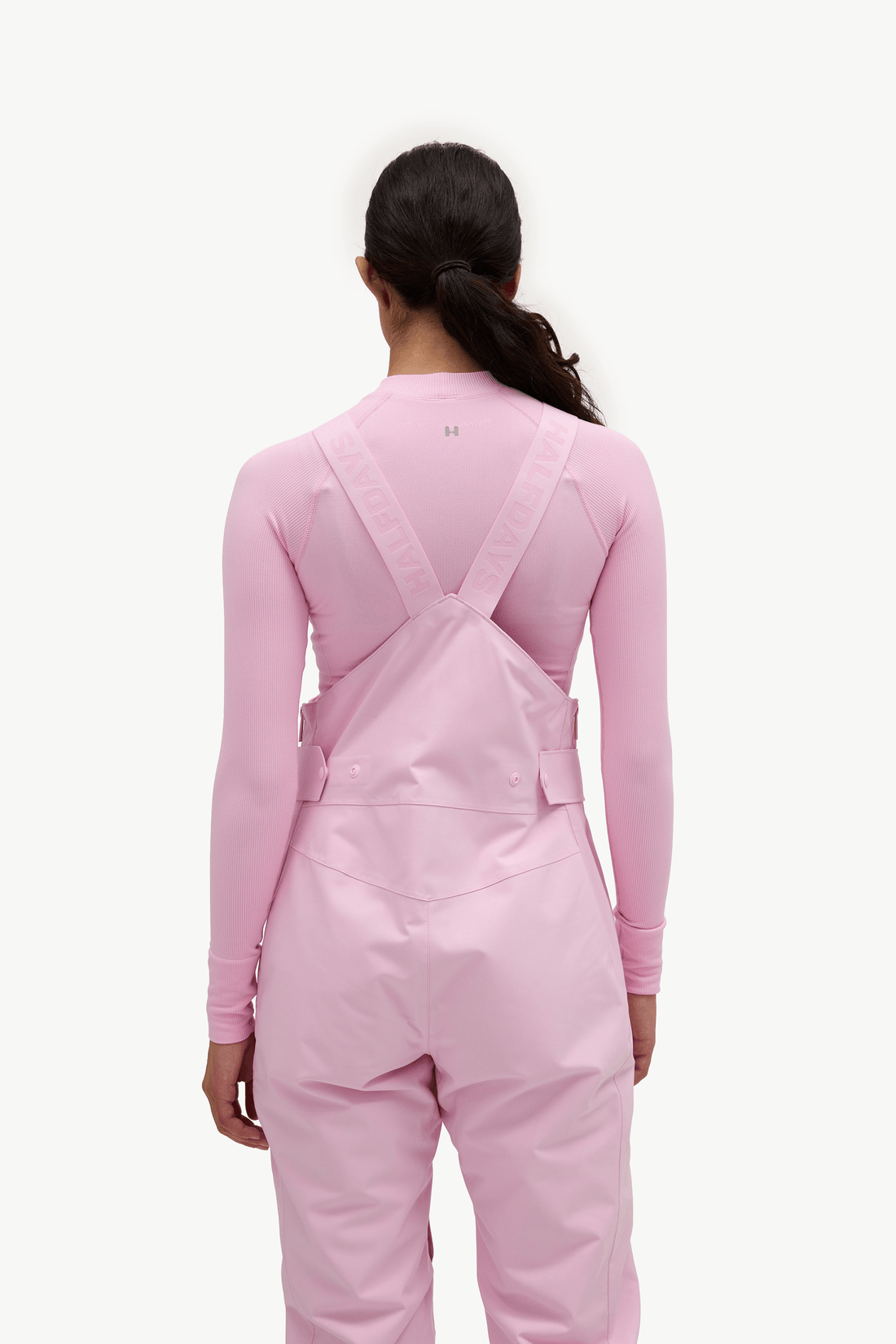 A model viewed from the back wearing a light pink long-sleeve top and matching pink bib overalls with adjustable shoulder straps, against a plain white background.