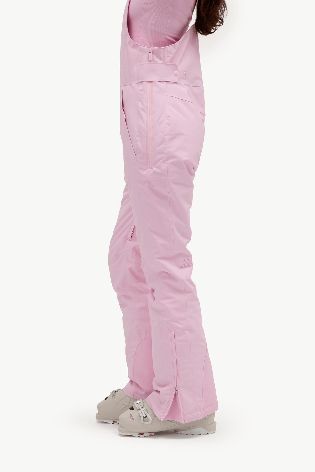 A side profile view of a person wearing light pink snow pants and beige ski boots against a plain white studio background.