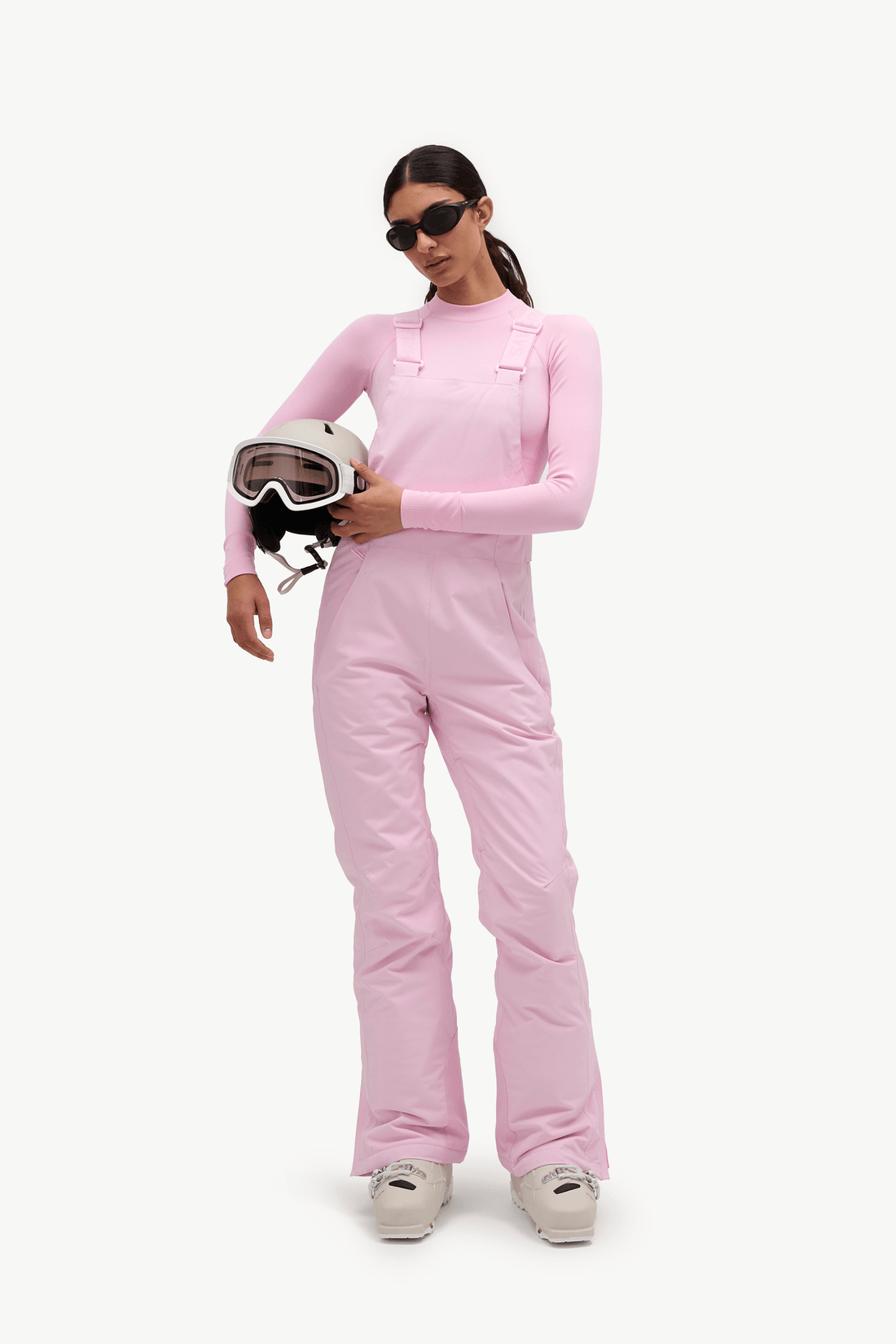 A model wearing a light pink ski jumpsuit and holding a white ski helmet with goggles, set against a plain white studio background.