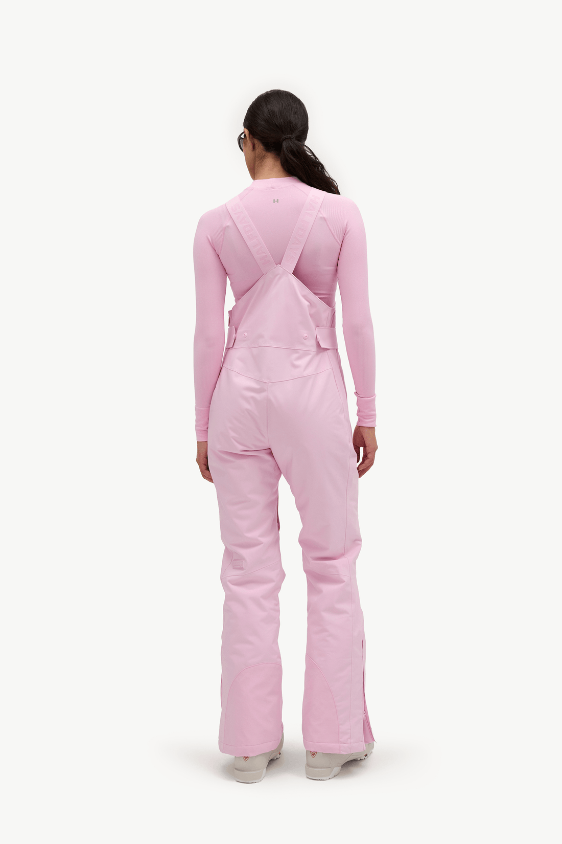 A model viewed from the back wearing a light pink long-sleeve jumpsuit with wide shoulder straps and flared legs, standing against a plain white studio background.