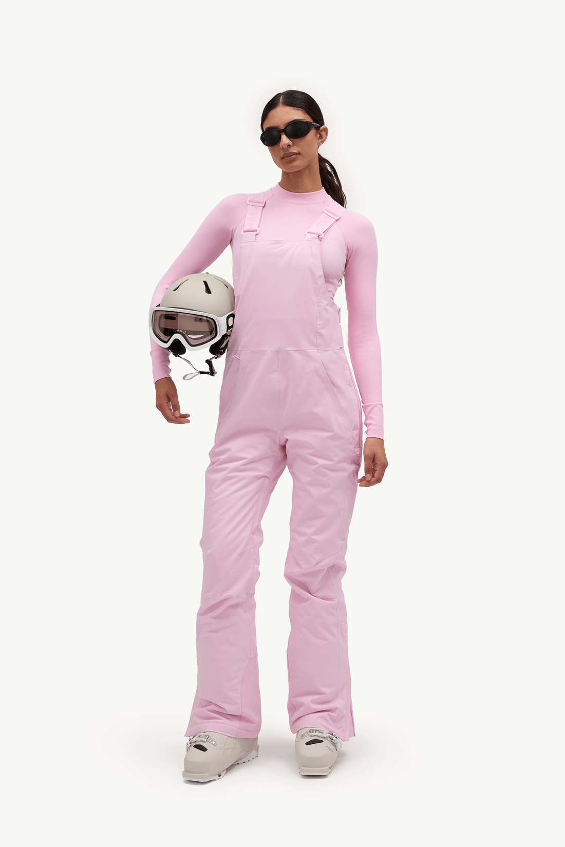 A model wearing a light pink ski jumpsuit and holding a white ski helmet, photographed against a clean white studio background.