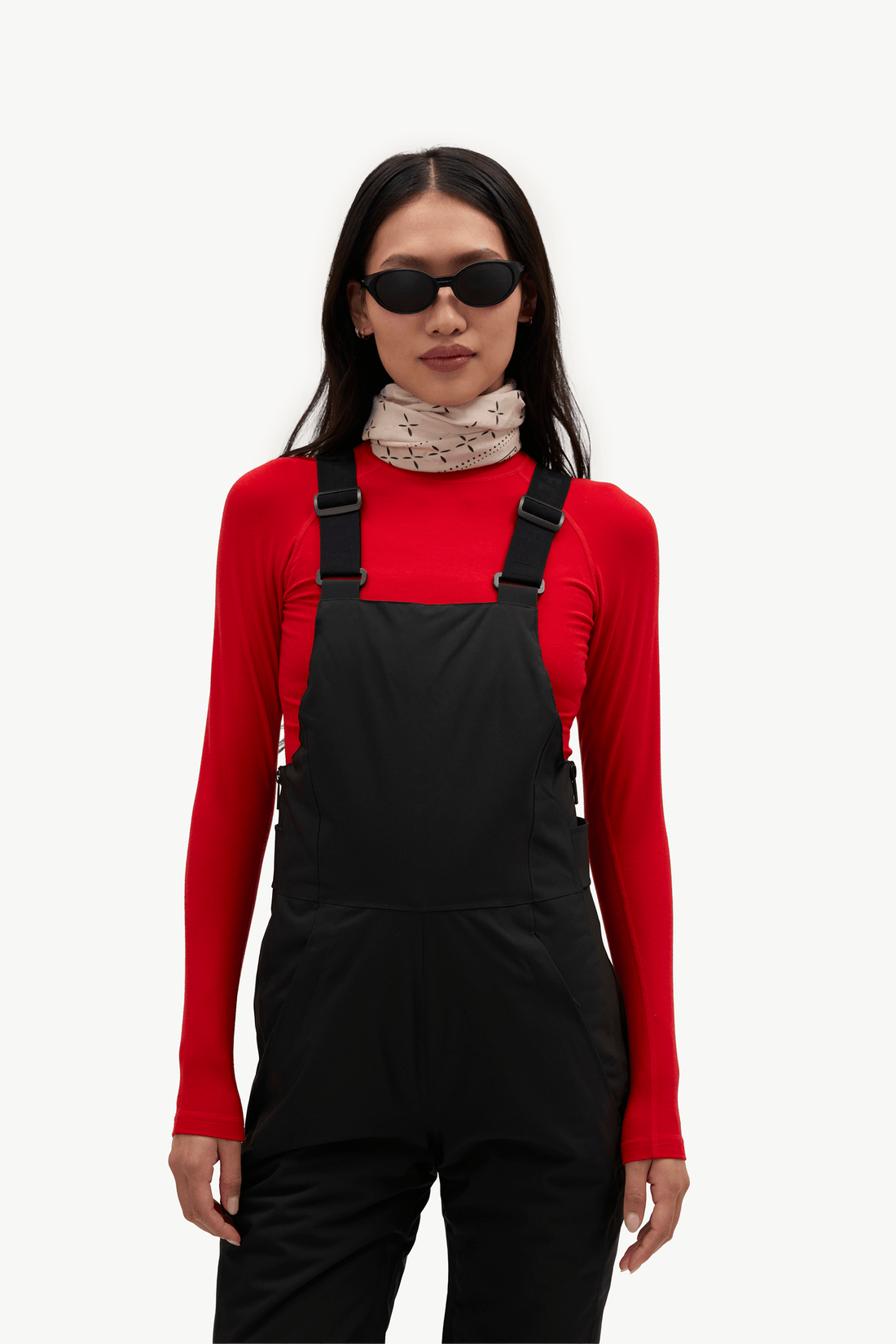 A model stands against a plain white studio background wearing black snow bib overalls over a bright red long-sleeve top, with a light patterned high-neck layer underneath.