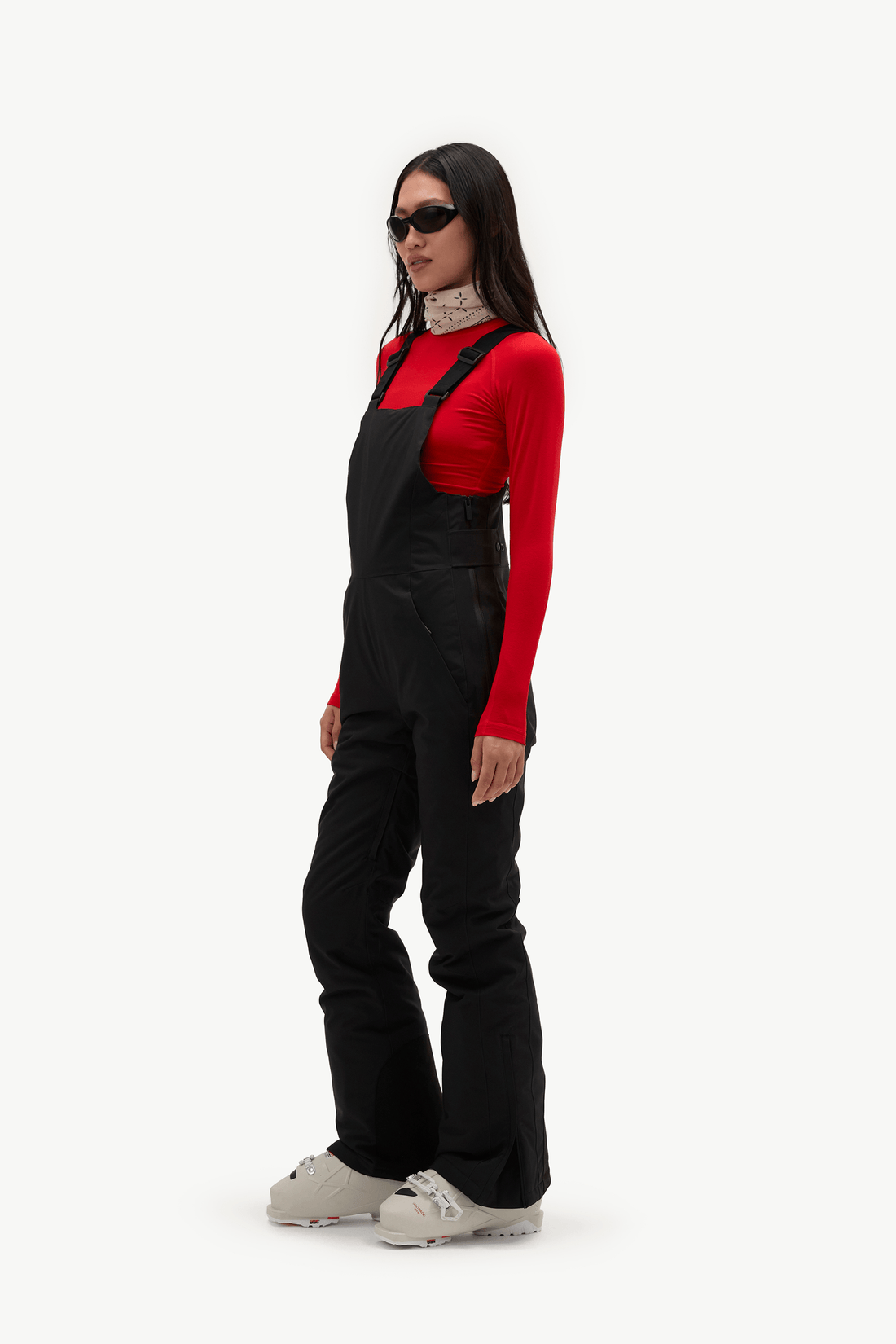 A model wearing black snow bib overalls over a red long-sleeve top and black sunglasses, standing against a plain white studio background.
