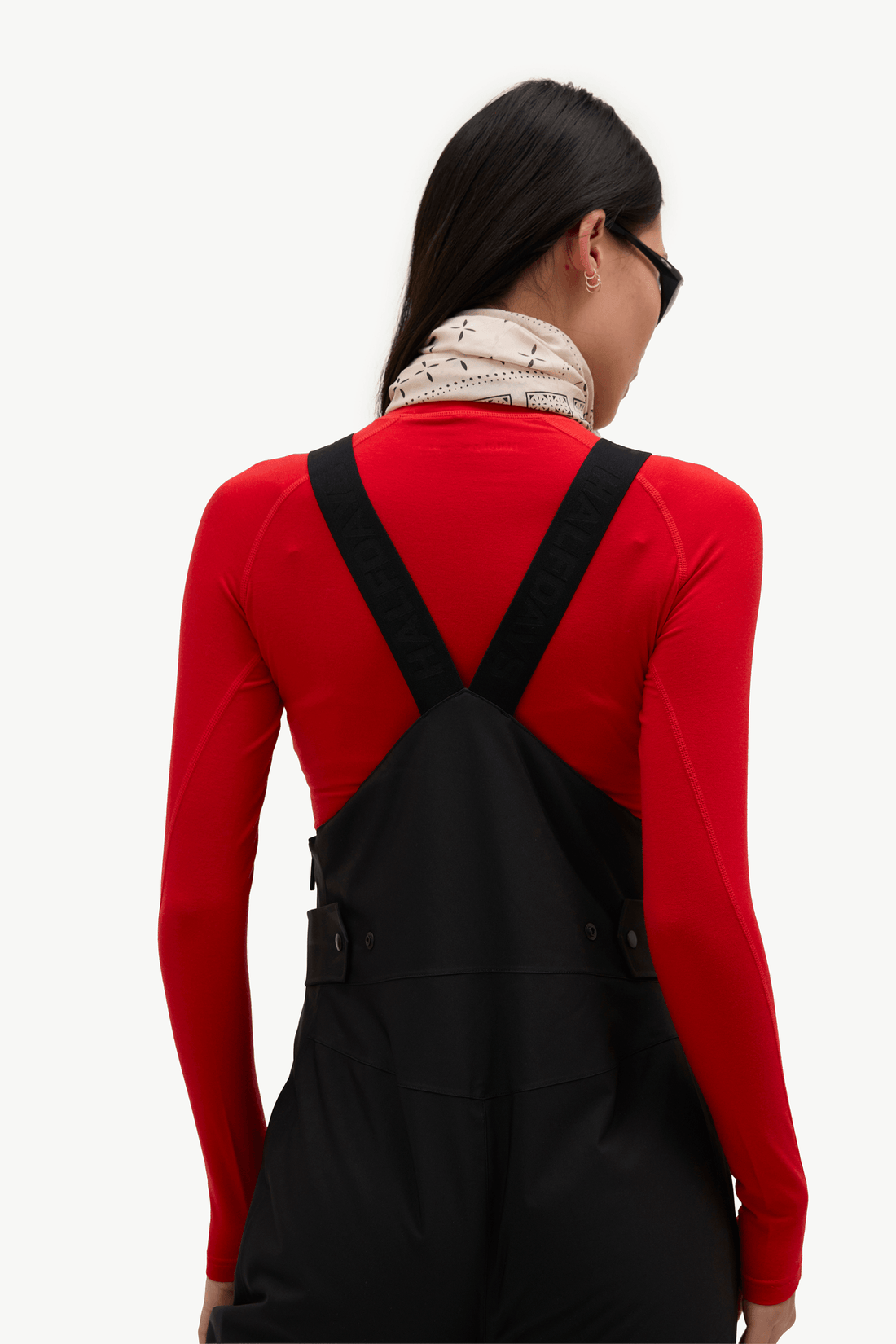 A woman shown from the back wearing a bright red long-sleeve top and black overall-style bib overalls with wide shoulder straps, against a plain light background.
