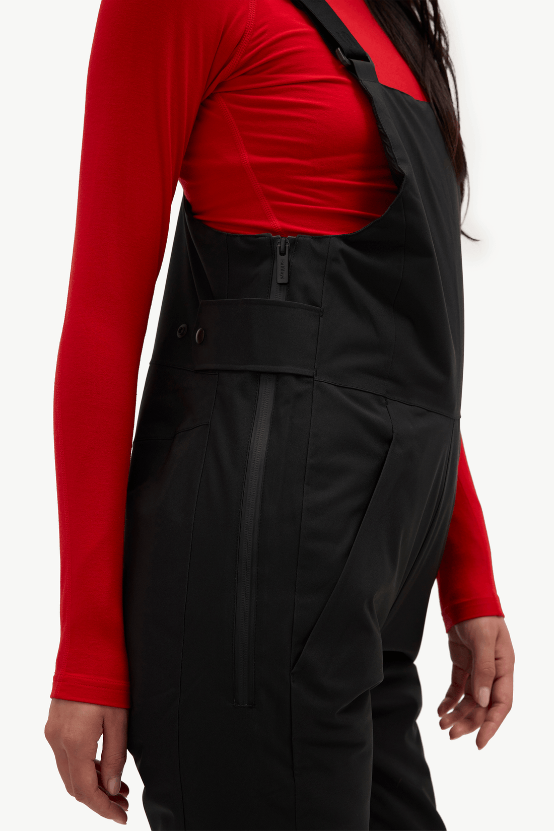 Black bib overalls worn by a model, styled over a bright red long-sleeve top, shown from the chest down against a plain white background.