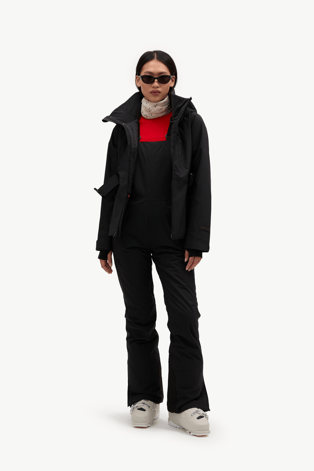 A model stands against a plain white studio background wearing black snow bib overalls over a bright red long-sleeve top, with a light patterned high-neck layer underneath and a black ski jacket. 
