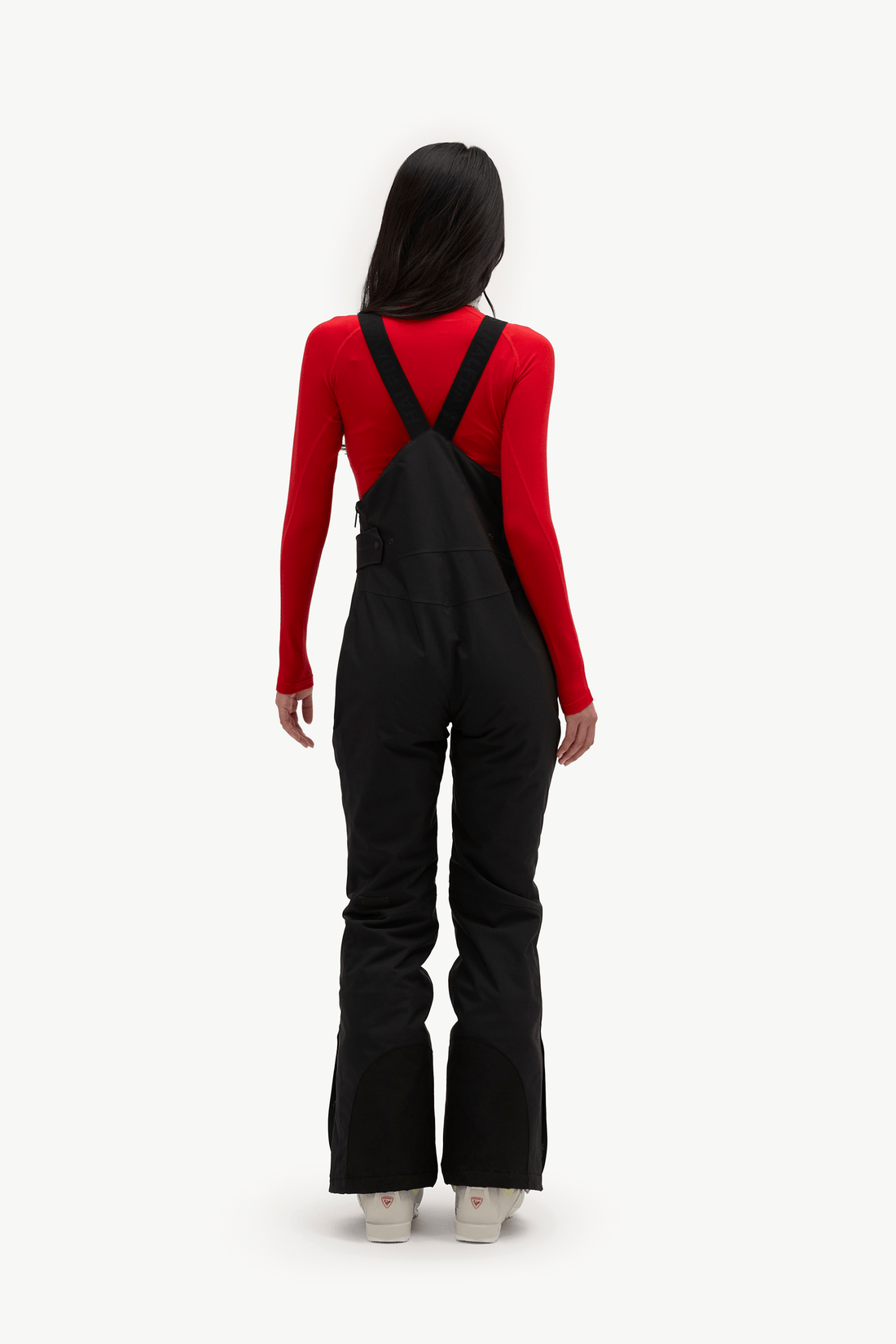 A model viewed from the back wearing black snowboarder overalls over a bright red long-sleeve top, standing against a solid white background.