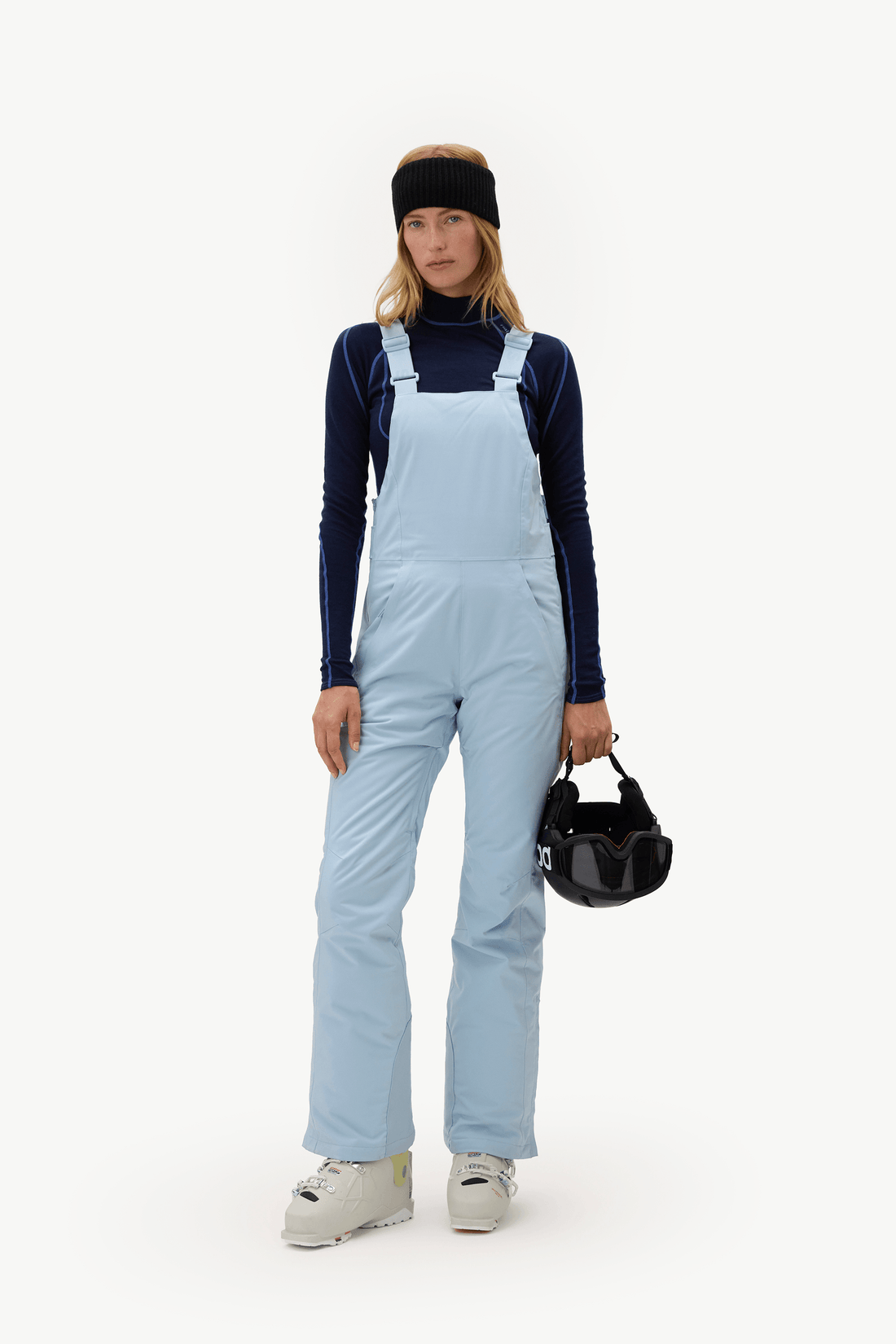 A model stands against a plain white studio background wearing light blue snow bib overalls over a dark navy long-sleeve top, with a black headband and white sneakers. She holds a small black handbag.