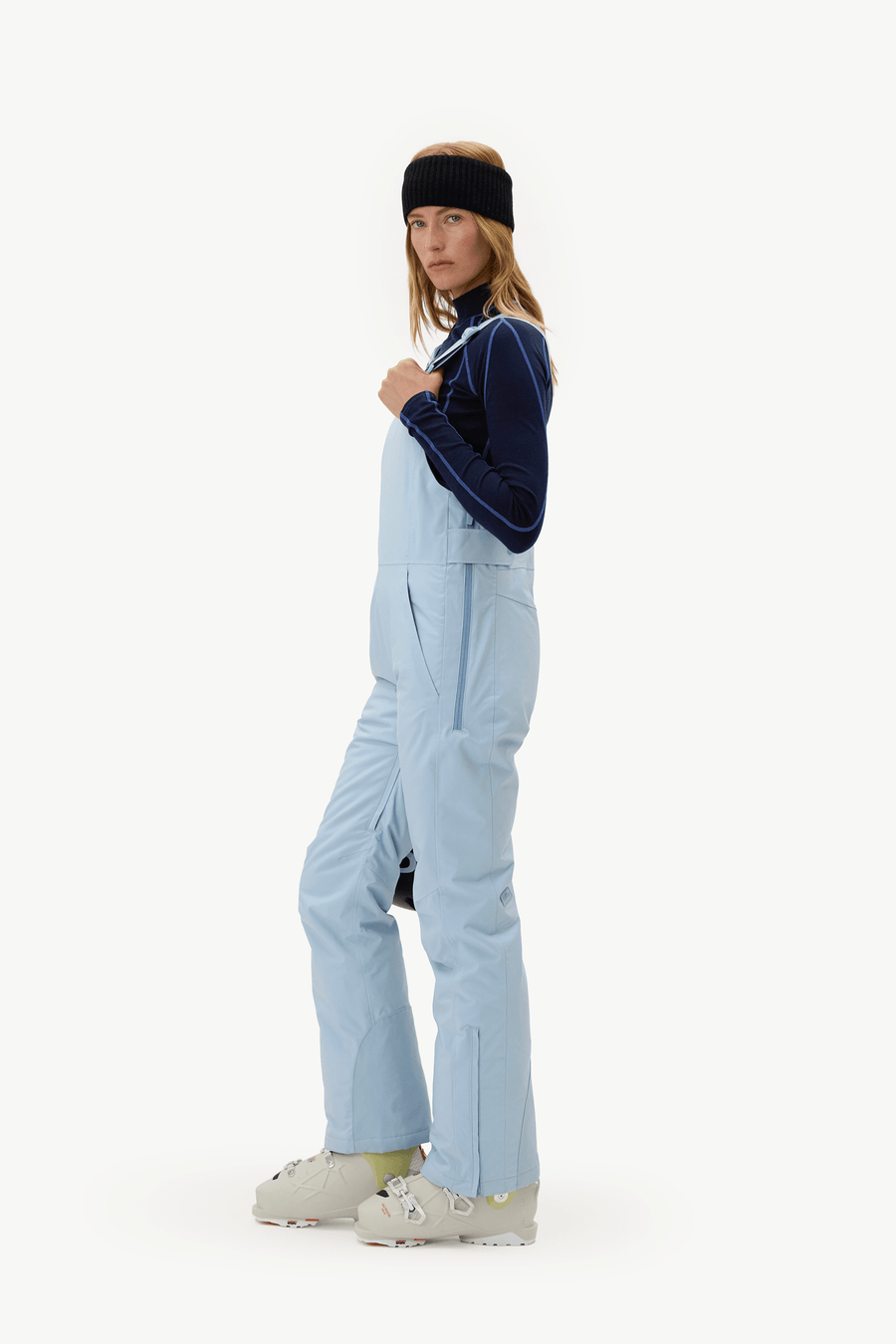 A model wearing light blue snow bib overalls, a navy blue long-sleeve top, a black headband, and ski boots, standing against a plain white studio background.