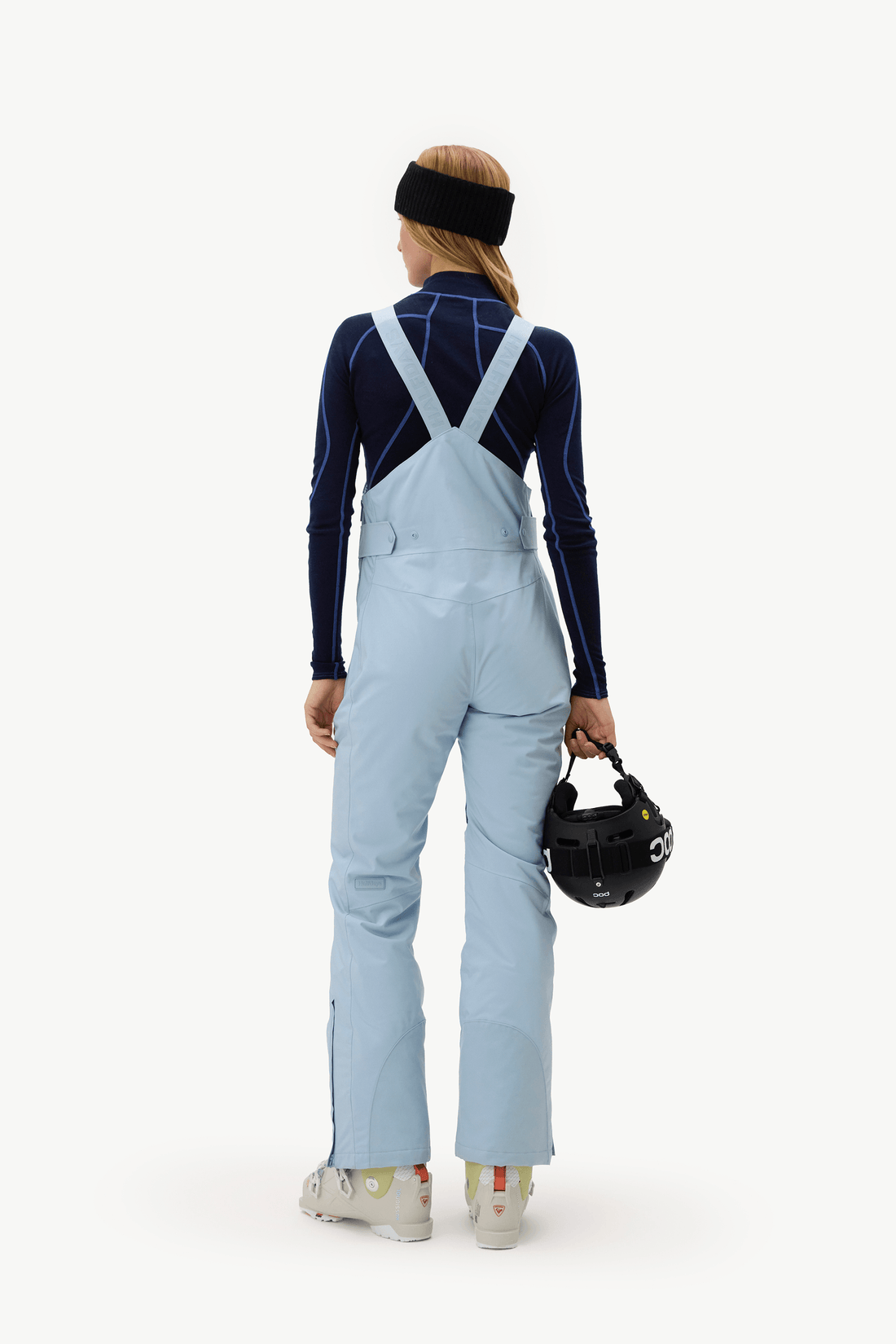 A woman shown from the back wearing light blue ski bib overalls over a dark navy long-sleeve athletic top, with a black headband and holding a black ski helmet against a plain white studio background.