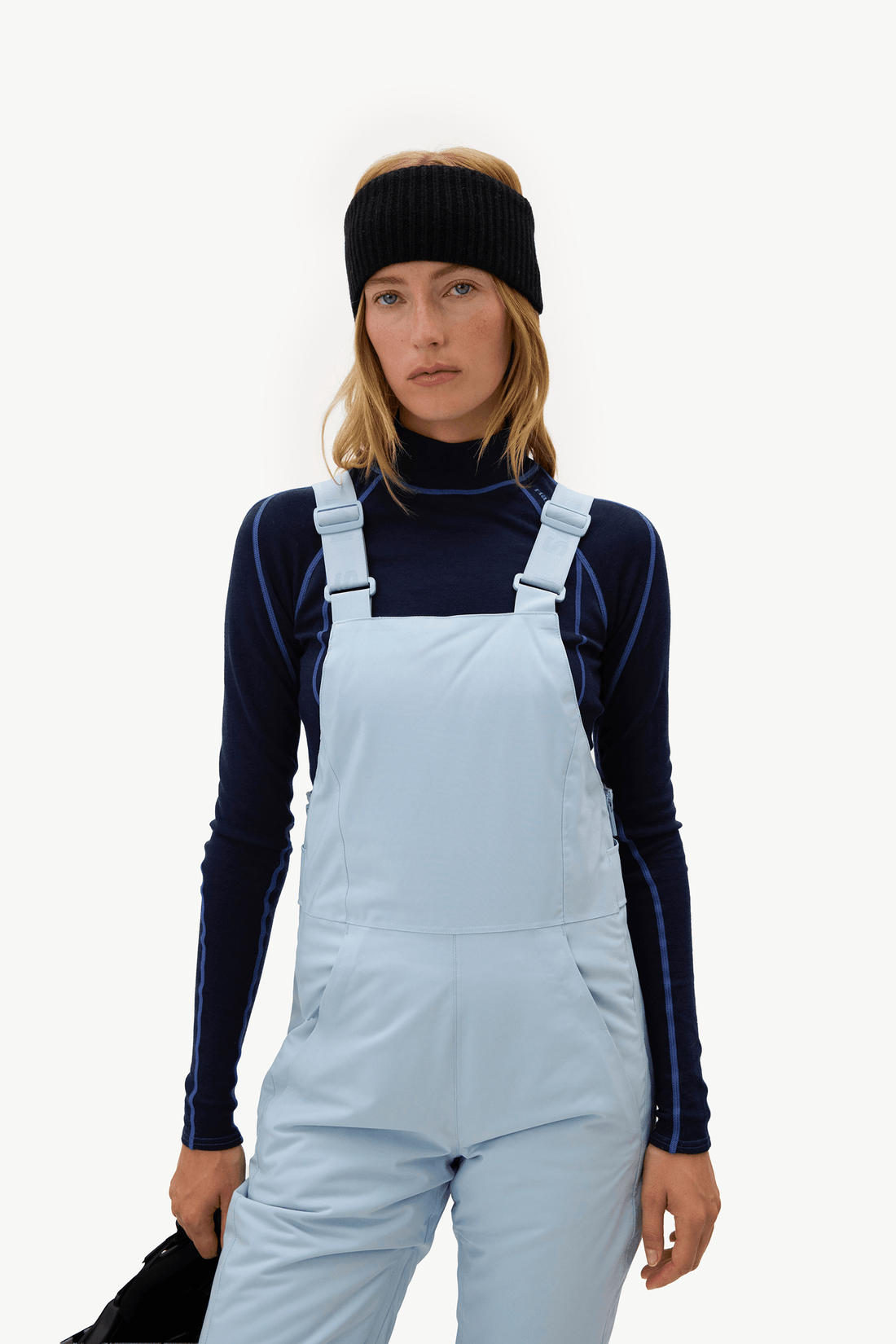 A woman wearing a black knit beanie, a navy long-sleeve turtleneck, and light blue ski overalls against a plain white studio background.