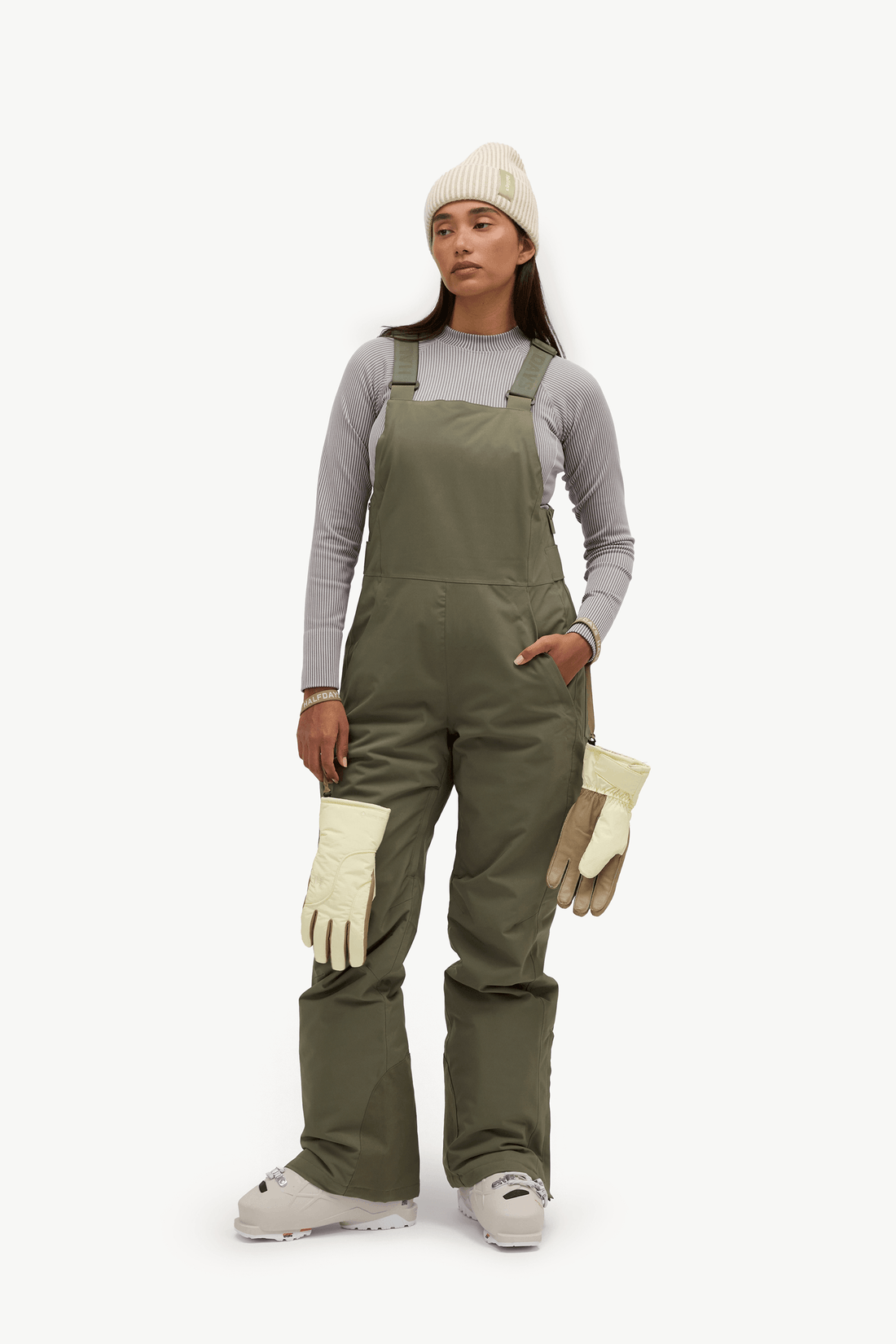 A model stands against a plain white studio background wearing olive green snow bib overalls over a light gray long-sleeve shirt, a cream knit beanie, off-white gloves, and light gray snow boots.