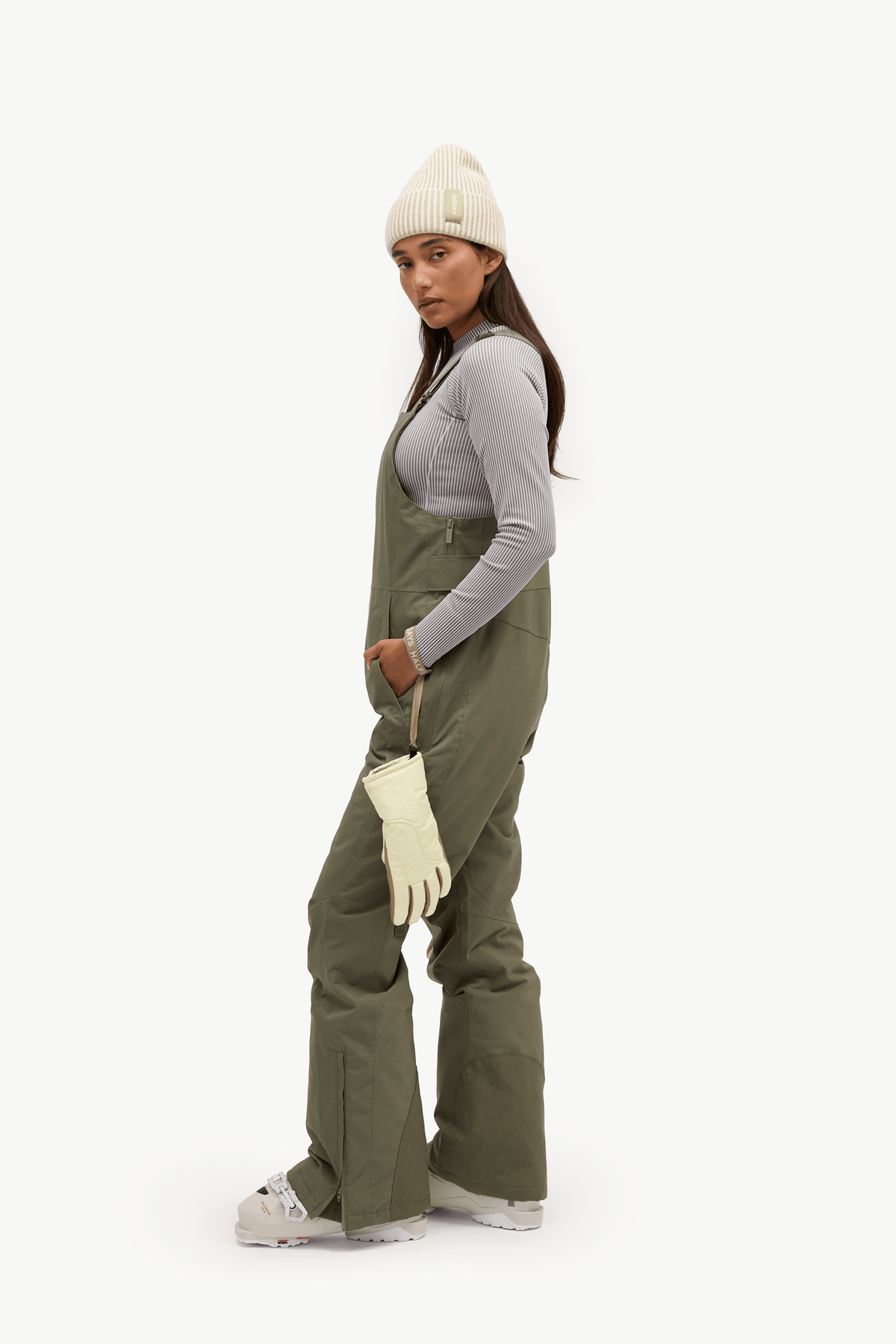 A woman modeling olive green snow bib overalls with a light gray long-sleeve top, cream beanie, cream gloves, and white sneakers, shown from a side profile against a plain white studio background.